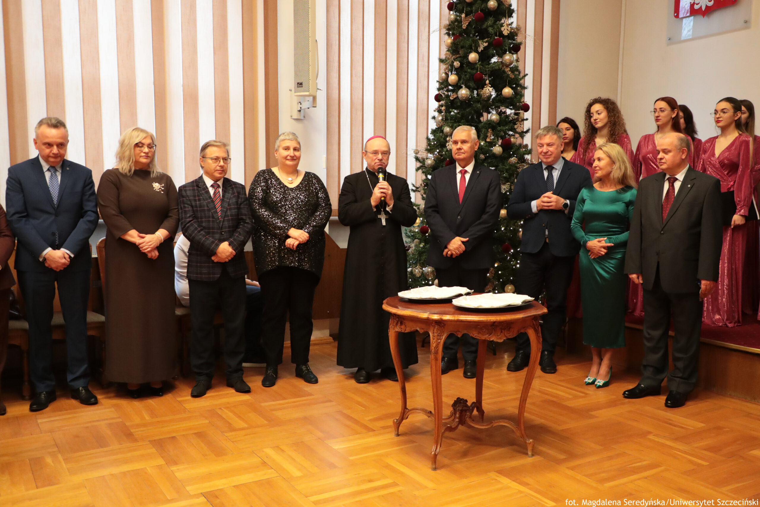 Grupa elegancko ubranych dorosłych osób stojących w pomieszczeniu obok udekorowanej choinki bożonarodzeniowej, z małym okrągłym stolikiem na pierwszym planie. W centrum stoi duchowny w czarnym stroju z krzyżem na piersi i trzyma mikrofon, jak podczas przemówienia. Po obu stronach stoją mężczyźni w garniturach oraz kobiety w wieczorowych sukienkach; po prawej stronie, na podwyższeniu, widać kilka młodych kobiet w długich czerwonych sukienkach, wyglądających jak chór. Choinka jest wysoka, ozdobiona bombkami w złotym i czerwonym kolorze oraz światełkami. Na stoliku leżą opłatki na srebrnych tacach. Tło stanowi ściana z pionowymi jasnymi i drewnianymi panelami; podłoga jest parkietowa. Na dole zdjęcia jest podpis fotografa i nazwa uczelni.