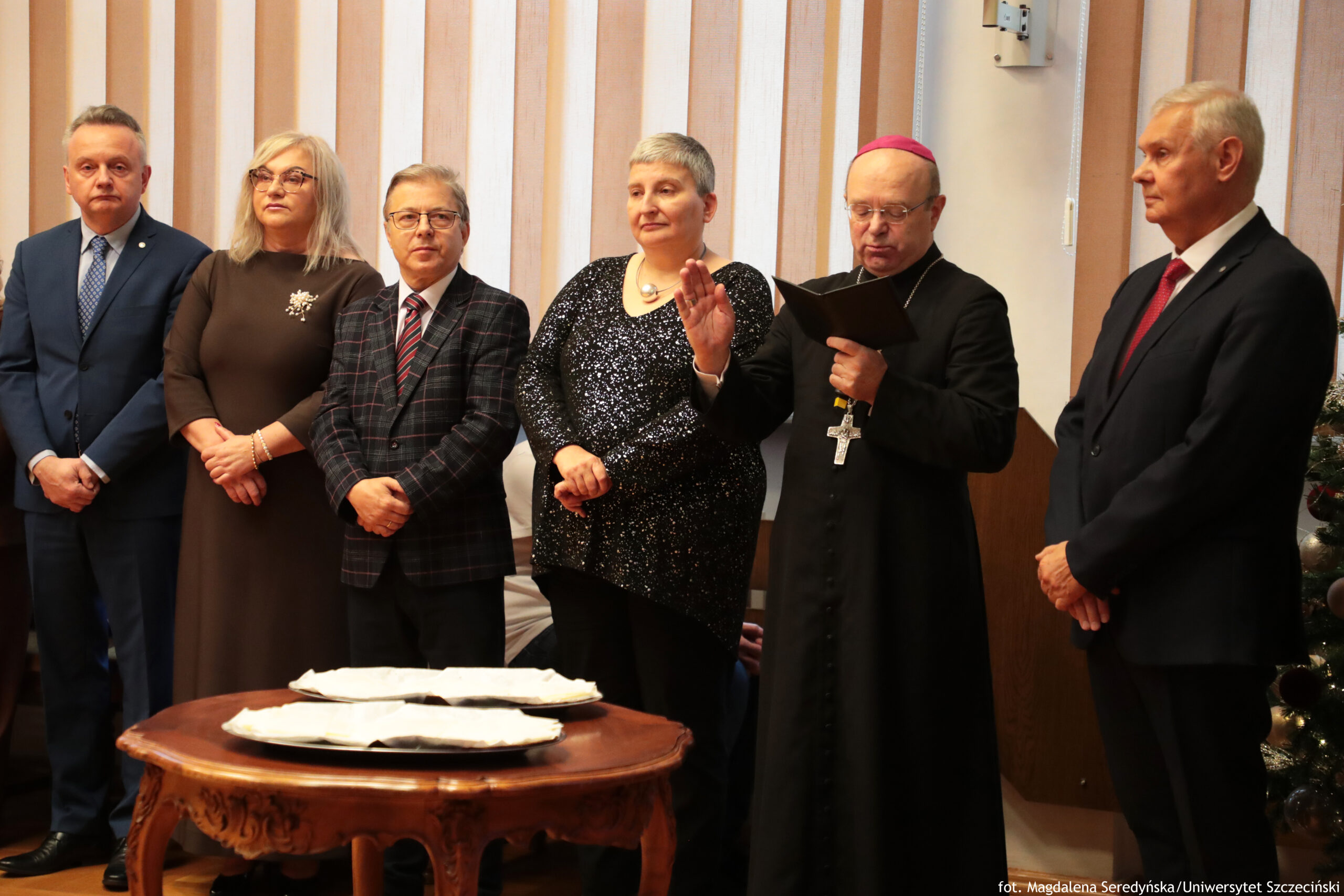 Grupa sześciu dorosłych osób stojących w pomieszczeniu przy okrągłym, rzeźbionym drewnianym stoliku z rozłożonymi opłatkami na tacach. To ujęcie wygląda jak uroczystość lub podpisanie/poświęcenie dokumentów. Od lewej stoi mężczyzna w granatowym garniturze i jasnej koszuli, z poważnym wyrazem twarzy. Obok niego kobieta o jasnych, blond włosach do ramion, w okularach, w długiej brązowej sukience z broszką na piersi. Trzecia osoba to mężczyzna w okularach, w marynarce w kratę i krawacie, z rękami złożonymi z przodu. Czwarta osoba to kobieta z krótkimi, siwymi włosami, w czarnej, błyszczącej sukience, stoi z rękami skrzyżowanymi. Piąta osoba to duchowny w czarnej szacie z dużym krzyżem na łańcuchu i różową piuską na głowie; trzyma otwartą książkę i ma uniesioną dłoń w geście błogosławieństwa lub przysięgi. Po prawej stronie stoi mężczyzna w ciemnym garniturze z czerwonym krawatem, zwrócony lekko w stronę duchownego. W tle są pionowe żaluzje w jasnych kolorach. Po prawej stronie częściowo widać choinkę. W prawym dolnym rogu jest podpis fotografa: „fot. Magdalena Seredyńska / Uniwersytet Szczeciński”.
