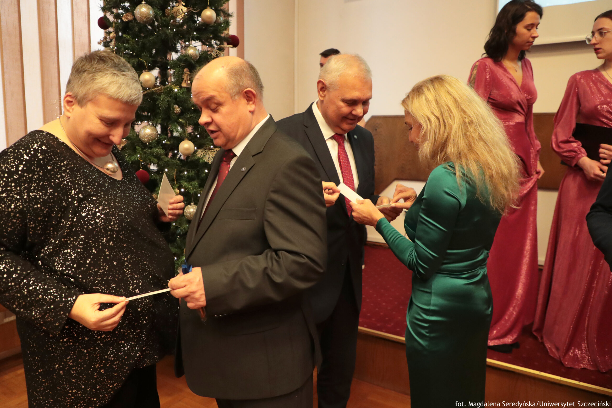 Grupa osób podczas uroczystości w sali przełamujący się opłatkiem, obok ubranej choinki. Na pierwszym planie po lewej jest kobieta z krótkimi siwymi włosami w czarnej, błyszczącej sukience, obok niej mężczyzna w ciemnym garniturze z czerwonym krawatem. Z tyłu stoi starszy mężczyzna w garniturze, uśmiecha się. Po prawej kobieta w dopasowanej, zielonej sukience trzyma opłatek i rozmawia z mężczyzną. W tle, na podwyższeniu, stoją dwie kobiety w długich, różowych sukienkach. Na dole po prawej widać mały napis: „fot. Magdalena Seredyńska/Uniwersytet Szczeciński”.