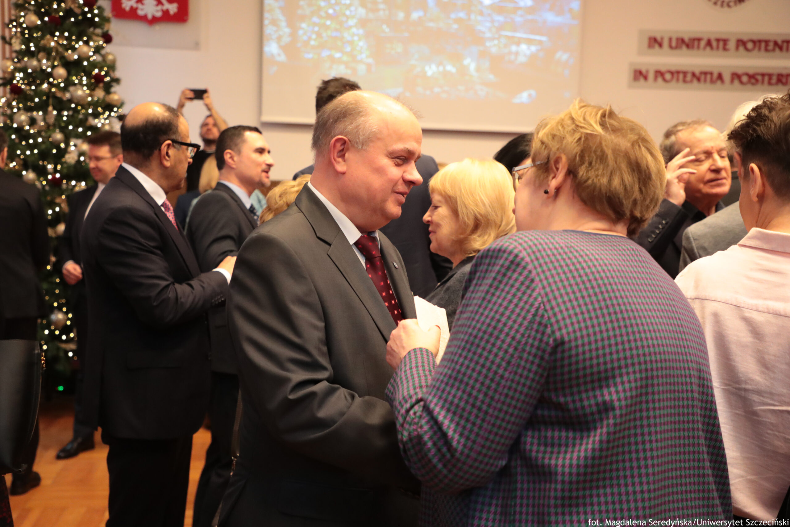 Grupa dorosłych osób podczas spotkania w sali, w tle widać dużą choinkę z lampkami. Na pierwszym planie mężczyzna w szarym garniturze i czerwonym krawacie rozmawia z kobietą w okularach, w granatowo‑zielonej sukience we wzór. Obok stoi blond kobieta, a w tle kilka osób w garniturach rozmawia w grupkach; jedna osoba trzyma telefon uniesiony do góry, jakby robiła zdjęcie. Z prawej strony na ścianie są fragmenty napisów po łacinie, a u dołu zdjęcia widoczny jest podpis: „fot. Magdalena Seredyńska/Uniwersytet Szczeciński”.