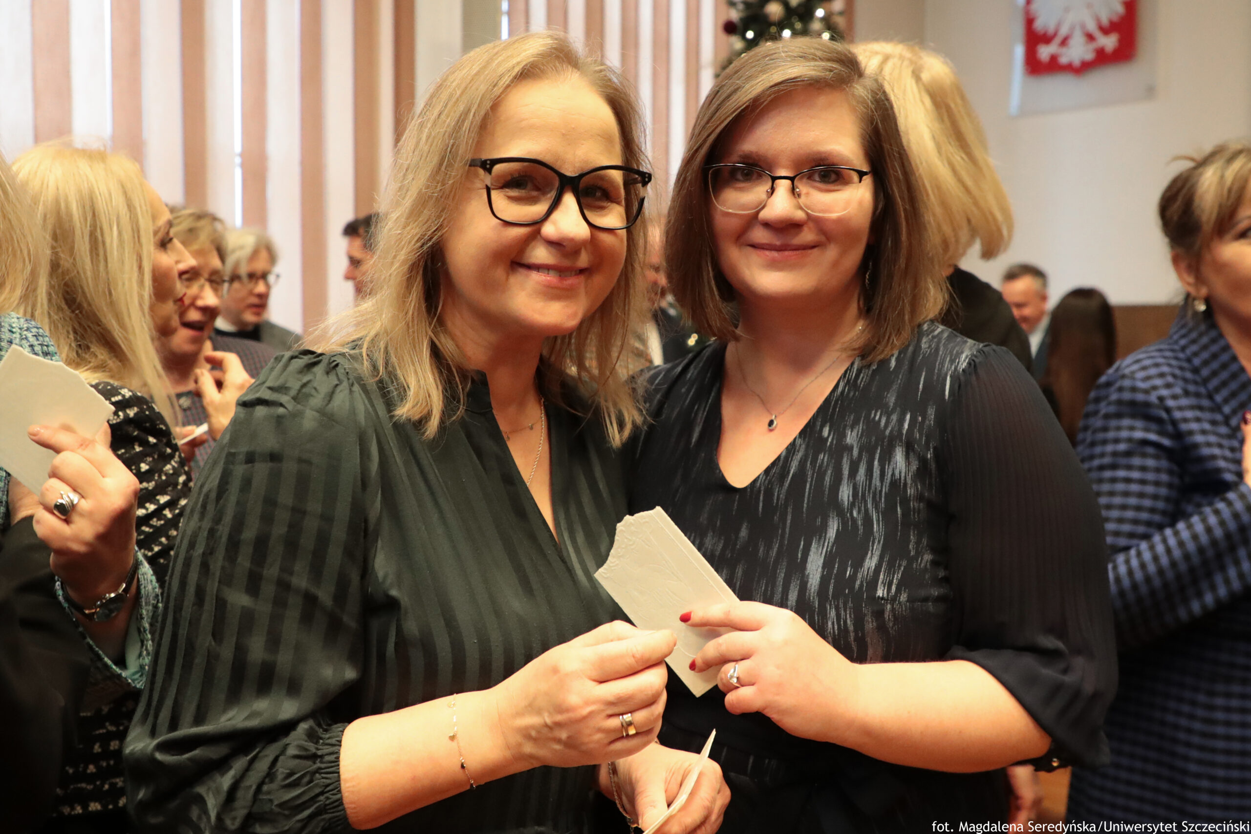 Dwie uśmiechnięte kobiety w średnim wieku stojące blisko siebie na pierwszym planie, w pomieszczeniu podczas spotkania. Obie mają jasną skórę i okulary; kobieta po lewej ma dłuższe, jasnoblond włosy i ciemnozieloną sukienkę z delikatnymi pionowymi paskami, a kobieta po prawej ma brązowe włosy do ramion i ciemną sukienkę z subtelnym, jaśniejszym wzorem. Trzymają w dłoniach opłatek. W tle widać kilka innych osób, pionowe żaluzje w oknach oraz fragment dekoracji świątecznej i czerwony element z białym orłem. W prawym dolnym rogu widoczny jest podpis: „fot. Magdalena Seredyńska / Uniwersytet Szczeciński”.