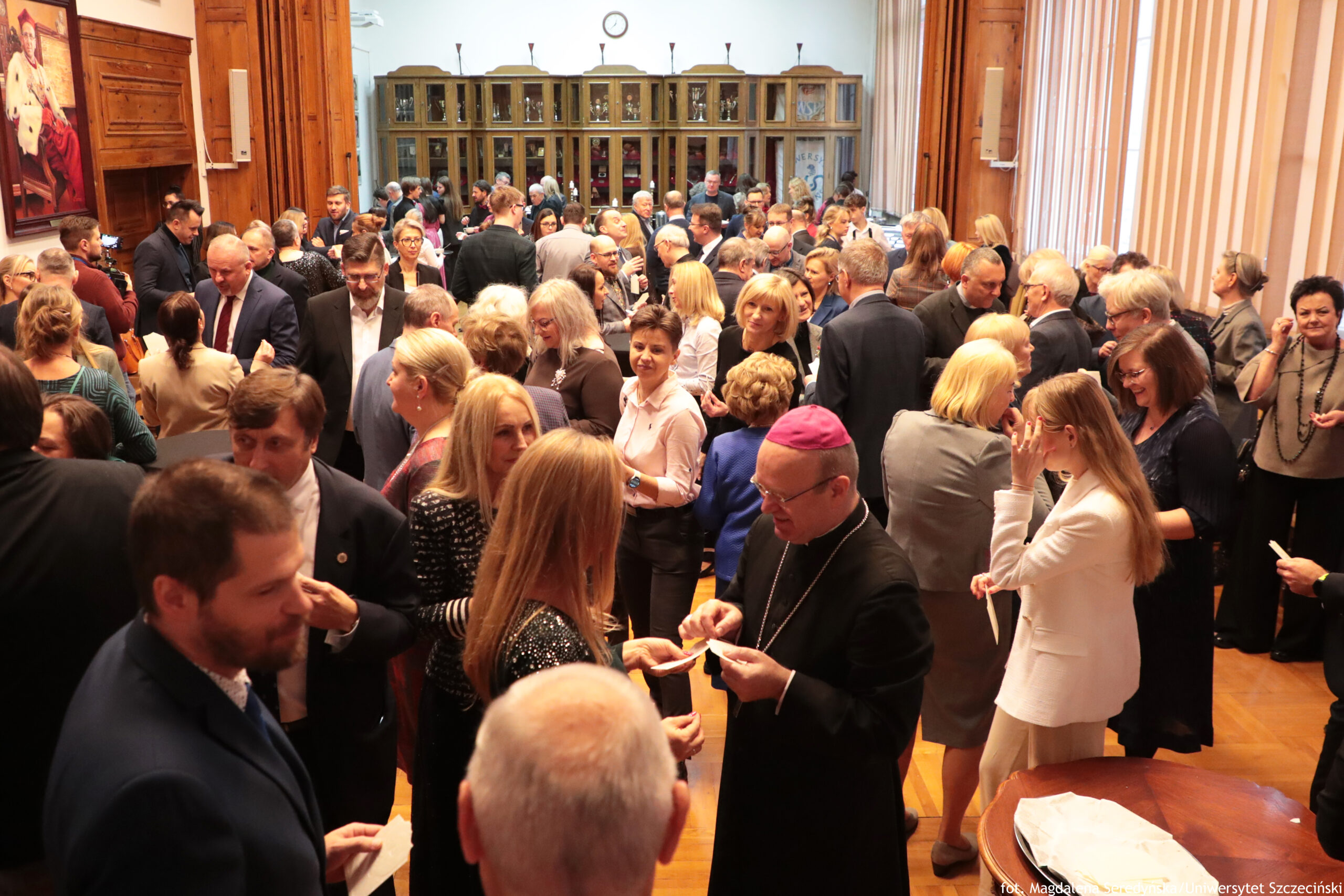 Zatłoczona sala z dużą liczbą dorosłych osób w strojach formalnych, stojących w grupach i rozmawiających. W pomieszczeniu widać drewniane ściany i podłogę oraz wysokie, przeszklone szafy w tle. Na pierwszym planie po prawej stronie znajduje się duchowny w czarnym stroju z różową piuską na głowie; trzyma w dłoniach małe kartki lub dokumenty. Wokół niego stoją kobiety i mężczyźni, część z nich również trzyma kartki. Po lewej stronie na ścianie wisi duży obraz przedstawiający siedzącą postać. W tle, nad szafami, wisi zegar ścienny.