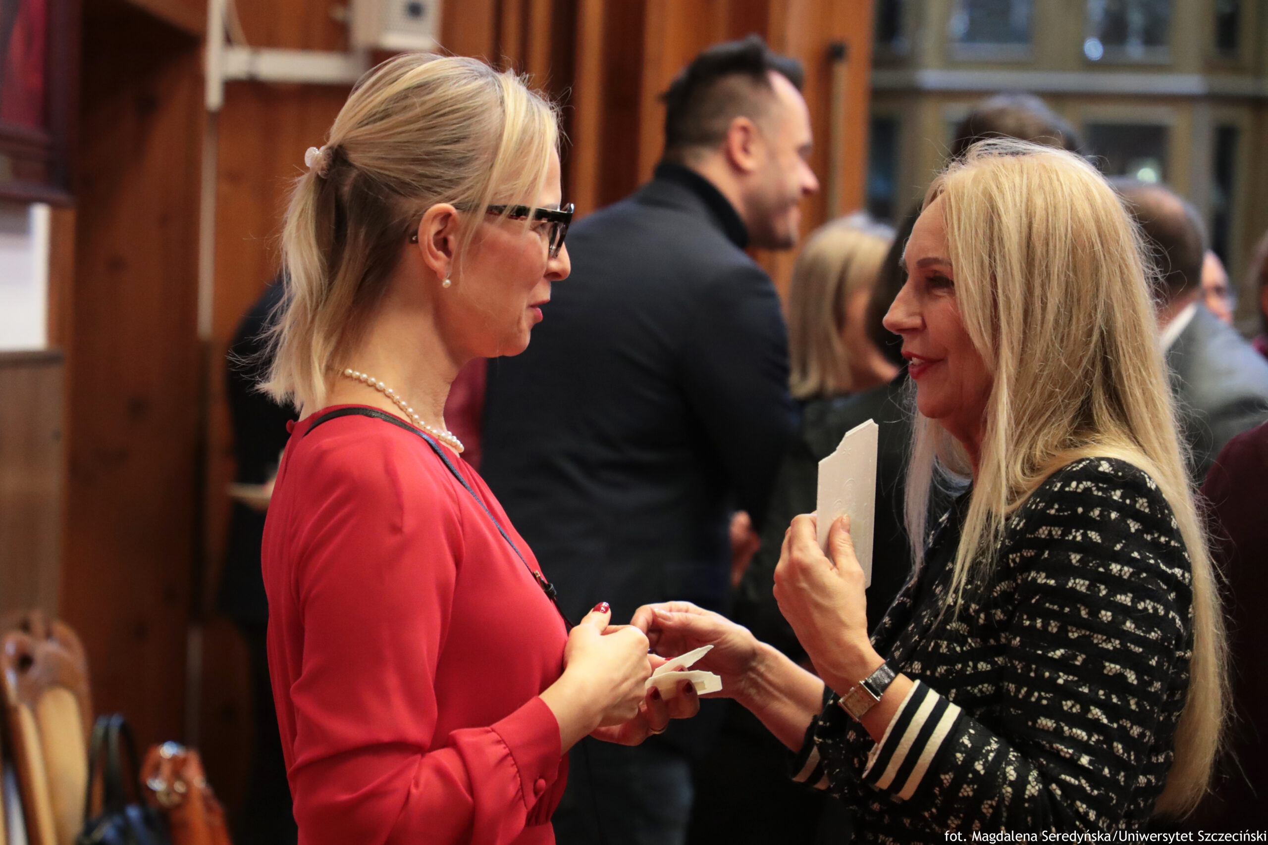 Dwie kobiety rozmawiające twarzą w twarz w pomieszczeniu o drewnianych ścianach. Kobieta po lewej ma jasne blond włosy związane z tyłu, okulary w czarnych oprawkach, kolczyk i naszyjnik z pereł; ma na sobie czerwoną sukienkę lub bluzkę i trzyma w dłoniach opłatek. Kobieta po prawej ma długie jasne blond włosy rozpuszczone, ma na sobie czarną marynarkę w jasny wzór oraz mankiety w biało-czarne paski; w jednej dłoni trzyma opłatek, a drugą wyciąga w stronę dłoni kobiety po lewej. W tle widać kilka innych osób w formalnych strojach, w tym mężczyznę w ciemnym ubraniu, oraz okna. W prawym dolnym rogu jest podpis: „fot. Magdalena Seredyńska/Uniwersytet Szczeciński”.