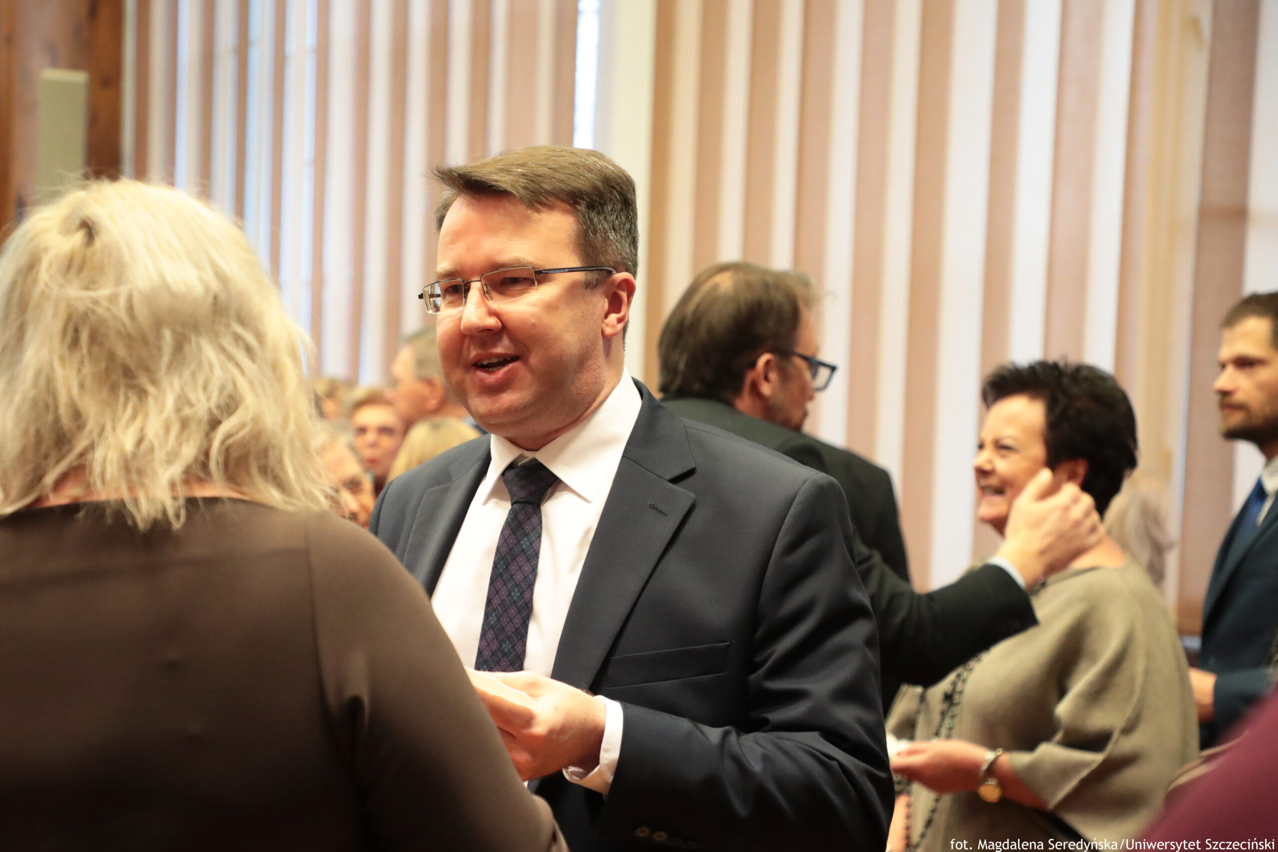 Grupa ludzi rozmawiających w pomieszczeniu z pionowymi żaluzjami w tle. Na pierwszym planie stoi mężczyzna w średnim wieku o jasnej karnacji, w okularach, z krótkimi brązowymi włosami, ubrany w granatowy garnitur, białą koszulę i ciemny krawat w kratę; rozmawia z osobą stojącą tyłem, z jasnymi, falowanymi włosami i w ciemnej bluzce. W tle widać kilka innych osób w eleganckich ubraniach, rozmawiających w grupkach; po prawej stronie uśmiechnięta kobieta z krótkimi ciemnymi włosami i jasną karnacją stoi obok mężczyzny w garniturze. W prawym dolnym rogu jest mały napis: „fot. Magdalena Seredyńska/Uniwersytet Szczeciński”.
