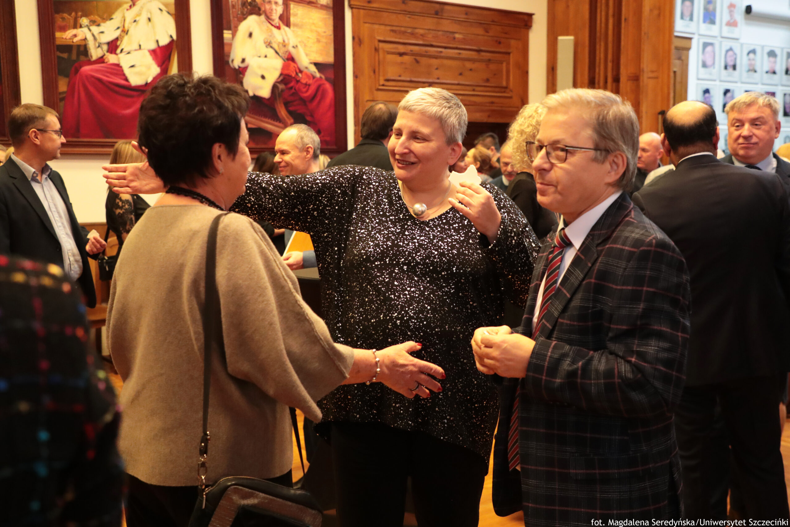 Grupa dorosłych osób rozmawiających w sali z drewnianymi elementami wystroju i dużymi obrazami na ścianach. W centrum stoi uśmiechnięta kobieta o krótkich siwych włosach, w czarnej bluzce w białe drobne wzory, z wyciągniętymi rękami w geście powitania lub obejmowania rozmówców. Po lewej jest kobieta odwrócona bokiem/tyłem, w beżowym okryciu i z czarną torebką na pasku. Po prawej stoi mężczyzna w okularach, w ciemnej kraciastej marynarce i krawacie w czerwono-granatowe pasy. W tle widać więcej osób w strojach formalnych oraz ścianę z wieloma małymi portretami. Na dole po prawej znajduje się podpis: „fot. Magdalena Seredyńska/Uniwersytet Szczeciński”.