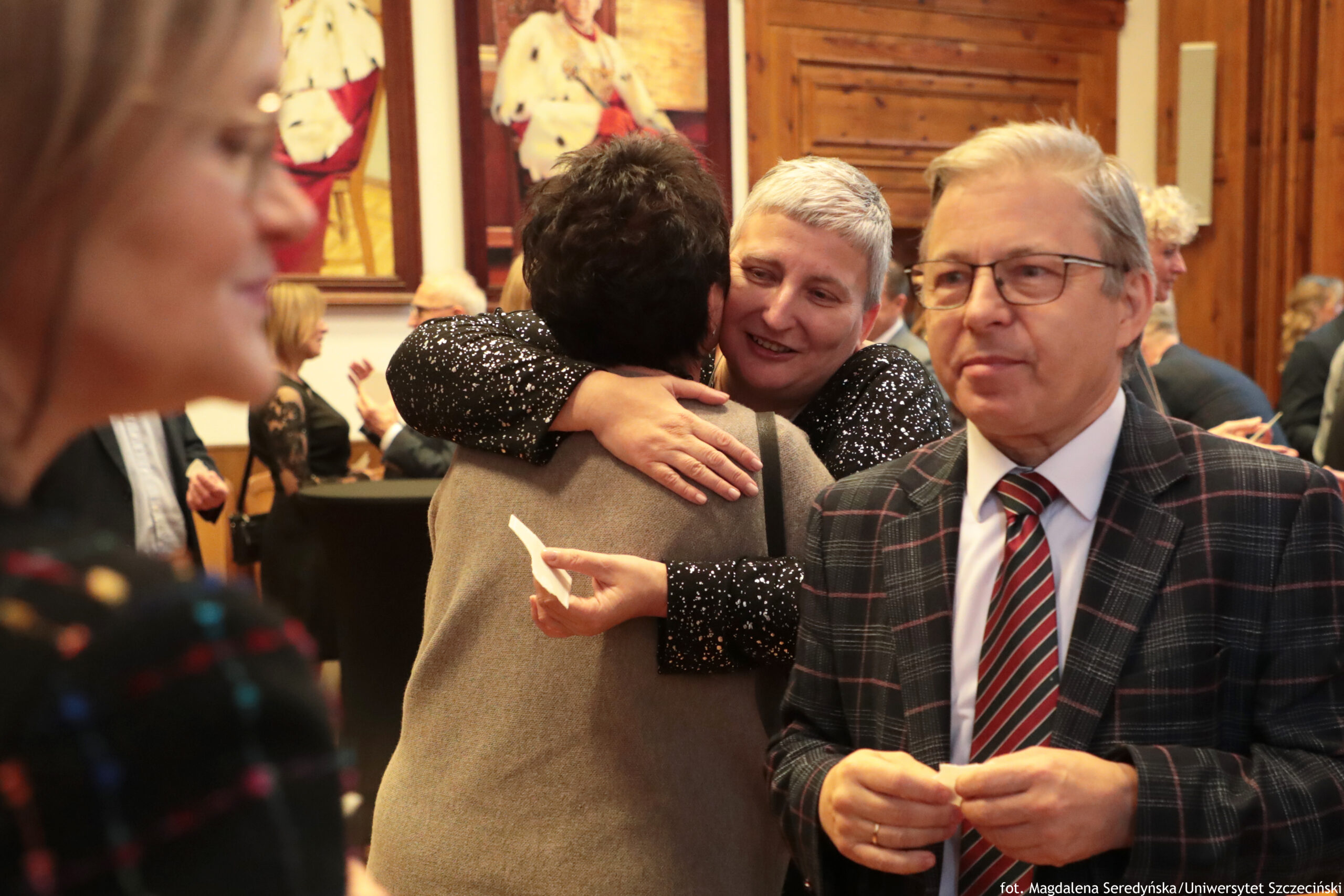 Grupa ludzi na wigilii, w sali z drewnianymi drzwiami i obrazami na ścianach. Na środku zdjęcia jasnowłosa kobieta o krótkich włosach przytula inną osobę w szarym swetrze; twarz przytulanej osoby jest odwrócona tyłem. Po prawej stronie stoi starszy mężczyzna w okularach, w kratkowanej marynarce i krawacie w czerwono-czarne pasy, trzyma w dłoniach mały przedmiot. Po lewej stronie, bardzo blisko aparatu, widać rozmyty profil kobiety w okularach. W tle są kolejne osoby rozmawiające przy wysokich stolikach. W prawym dolnym rogu jest podpis: „fot. Magdalena Seredyńska/Uniwersytet Szczeciński”.