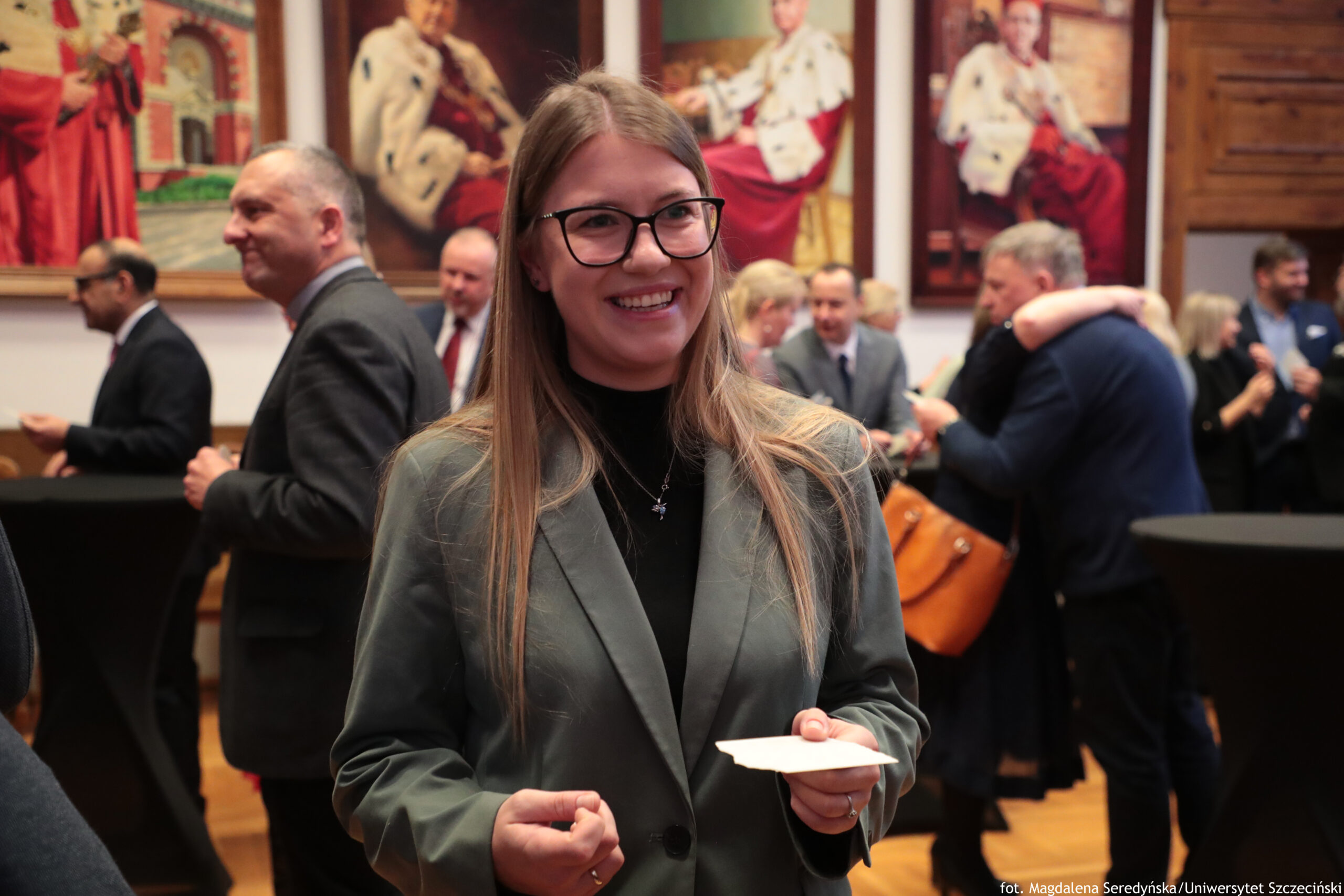 Uśmiechnięta kobieta w okularach, z długimi jasnymi włosami, ubrana w ciemny golf i zielonkawą marynarkę, trzyma w dłoniach opłatek. Ma jasną karnację. Scena wygląda na wydarzenie towarzyskie lub oficjalne w pomieszczeniu. W tle stoi kilka osób w garniturach i eleganckich strojach, rozmawiają w grupkach; po prawej widać parę obejmującą się. Na ścianach wiszą duże obrazy przedstawiające postacie w ceremonialnych szatach. Na dole po prawej jest podpis: „fot. Magdalena Seredyńska/Uniwersytet Szczeciński”.