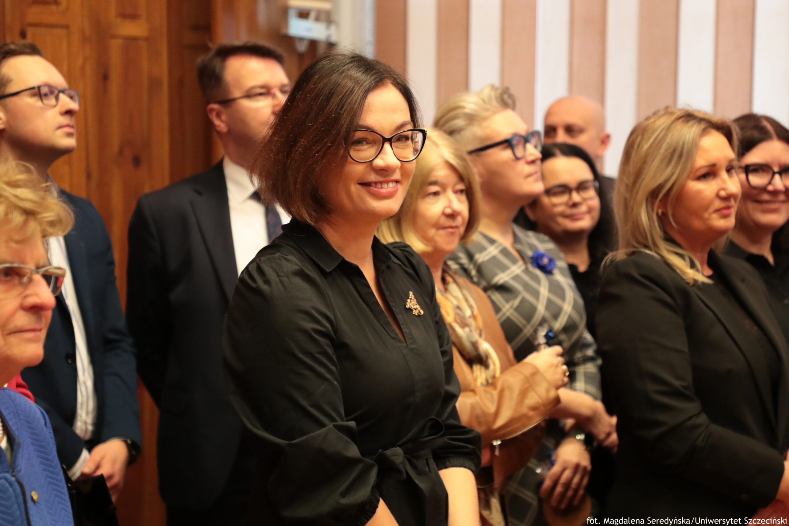 Grupa dorosłych osób stojących blisko siebie w pomieszczeniu, jak na formalnym spotkaniu. W centrum kadru znajduje się uśmiechnięta kobieta o jasnej skórze, krótkich ciemnobrązowych włosach do brody i w czarnych okularach; ma na sobie czarną sukienkę lub koszulową kreację z paskiem w talii i małą broszką przypiętą do piersi. Wokół niej stoją inne osoby w strojach biznesowych: mężczyźni w garniturach i kobiety w eleganckich ubraniach, część z nich w okularach. Tło tworzą drewniane drzwi lub boazeria po lewej oraz jasna ściana w pionowe pasy po prawej. W prawym dolnym rogu jest podpis: „fot. Magdalena Seredyńska/Uniwersytet Szczeciński”.
