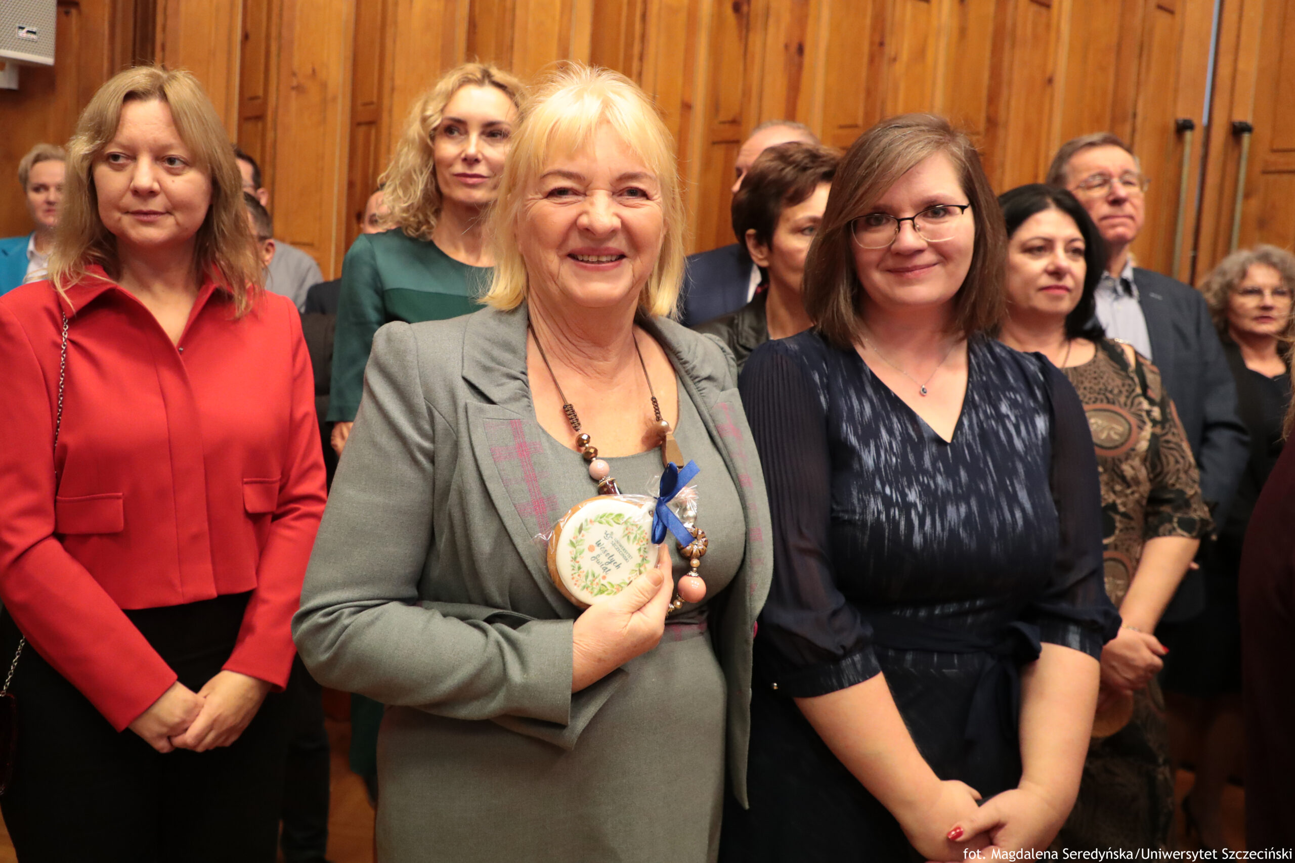 Grupa dorosłych osób stojących w pomieszczeniu z drewnianą, pionowo wyłożoną boazerią na ścianie. Na pierwszym planie w centrum stoi starsza kobieta o jasnej skórze i blond włosach do ramion, uśmiechnięta, w szarym kostiumie; na szyi ma długi naszyjnik i trzyma okrągły medal lub pamiątkę z jasną wstążką. Po lewej stronie stoi kobieta o jasnej skórze, w czerwonej koszuli, z jasnobrązowymi włosami do ramion, z neutralnym wyrazem twarzy. Po prawej stoi młodsza kobieta o jasnej skórze, w granatowej sukience, w okularach, z brązowymi włosami do ramion, lekko się uśmiecha. W tle widać kilka innych osób, kobiety i mężczyzn, częściowo zasłoniętych. W prawym dolnym rogu znajduje się podpis: „fot. Magdalena Seredyńska/Uniwersytet Szczeciński”.