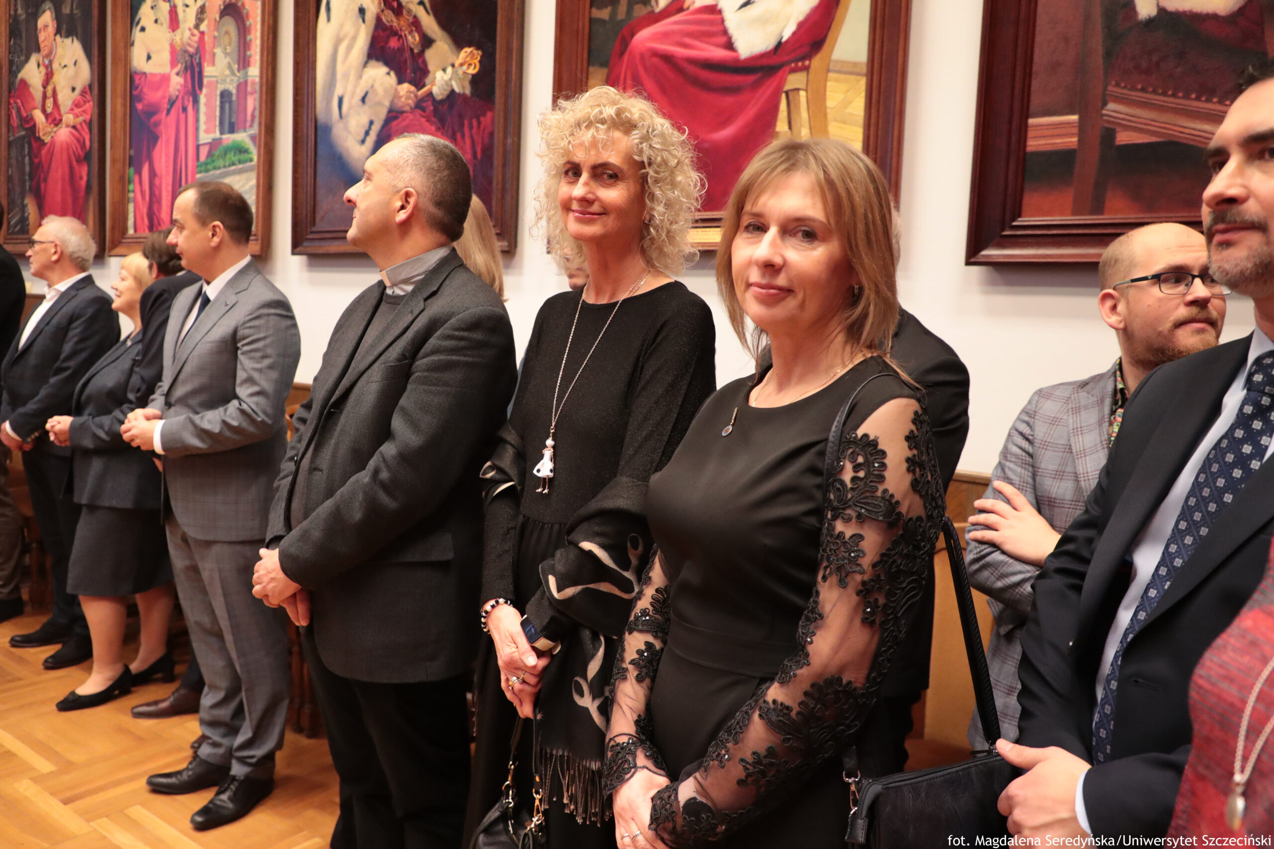 Grupa elegancko ubranych dorosłych osób stojących w pomieszczeniu, na tle dużych obrazów w ramach. W centrum kadru stoją dwie kobiety: jedna z jasnymi, kręconymi włosami w czarnej sukni i z długim naszyjnikiem, druga z prostymi, jasnobrązowymi włosami w czarnej sukience z przezroczystymi rękawami ozdobionymi czarną koronką. Wokół nich stoją mężczyźni w garniturach; po prawej widać też mężczyznę w okularach i w marynarce w kratę. Większość osób ma jasną karnację. Na ścianie za nimi wiszą duże portrety przedstawiające postacie w czerwonych strojach z białym futrem, przypominające królewskie lub ceremonialne szaty. W prawym dolnym rogu jest mały napis: „fot. Magdalena Seredyńska/Uniwersytet Szczeciński”.
