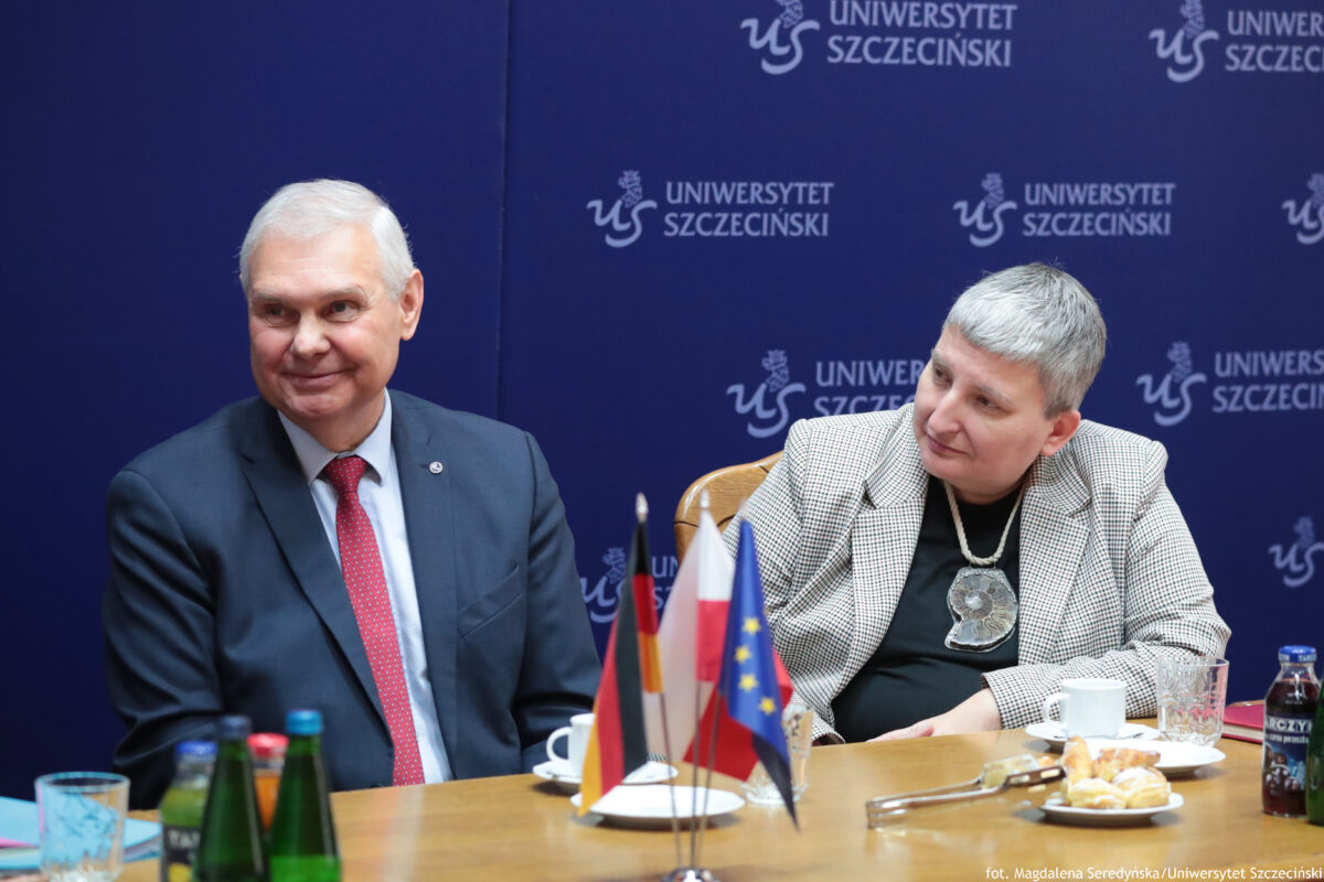 Dwójka osób siedzących przy stole podczas oficjalnego spotkania, na tle granatowej ścianki z powtarzającym się napisem „UNIWERSYTET SZCZECIŃSKI” i logo. Po lewej stronie siedzi mężczyzna o jasnej skórze i krótkich, białych włosach, w granatowym garniturze, białej koszuli i czerwonym krawacie w drobny wzór. Ma lekki uśmiech i jest zwrócony trochę w stronę prawej. Po prawej stronie siedzi kobieta o jasnej skórze i krótkich, siwych włosach, w jasnej, wzorzystej marynarce i czarnym ubraniu pod spodem. Na szyi ma duży, okrągły, metaliczny medalion na łańcuchu. Patrzy w stronę mężczyzny po lewej. Na stole stoją małe flagi na wspólnym stojaku: widać flagę Polski, flagę Unii Europejskiej i dwie inne (kolory czarno-czerwono-żółty oraz czerwono-biały). Na stole są też filiżanki, talerzyk z małymi wypiekami, sztućce, szklanki oraz kilka butelek z napojami. W prawym dolnym rogu widoczny podpis: „fot. Magdalena Seredyńska/Uniwersytet Szczeciński”.