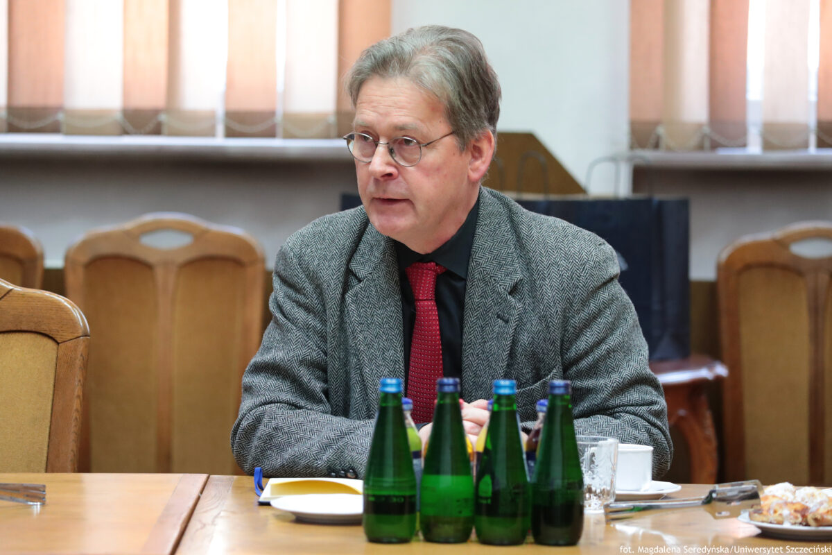 Mężczyzna w średnim lub starszym wieku siedzący przy stole w małej sali senatu. Ma jasną skórę, siwe włosy zaczesane na bok i okrągłe okulary. Jest ubrany w szarą marynarkę z drobnym wzorem, czarną koszulę i czerwony krawat. Przed nim na stole stoją cztery zielone szklane butelki z niebieskimi zakrętkami; obok leżą talerzyk, filiżanka, szklanka i sztućce, a z prawej strony widać talerz z ciastkami. W tle są drewniane krzesła oraz okno z pionowymi żaluzjami. W prawym dolnym rogu widnieje napis: „fot. Magdalena Seredyńska/Uniwersytet Szczeciński”.