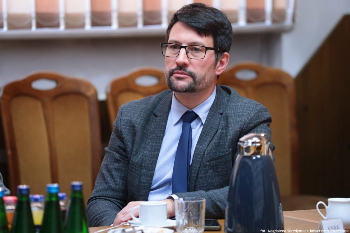 Mężczyzna w garniturze siedzący przy stole w małej sali senatu. Ma jasną karnację, krótkie ciemne włosy, okulary w czarnych oprawkach oraz krótko przystrzyżoną brodę i wąsy. Ubrany jest w szarą marynarkę w kratę, jasnoniebieską koszulę i ciemnoniebieski krawat; ma poważny, skupiony wyraz twarzy i patrzy lekko w bok. Na stole stoją filiżanki, szklanka, kilka butelek napojów i duży ciemny dzbanek/termos z metaliczną pokrywką. W tle znajdują się rzędy tapicerowanych krzeseł. W prawym dolnym rogu widoczny jest mały napis.