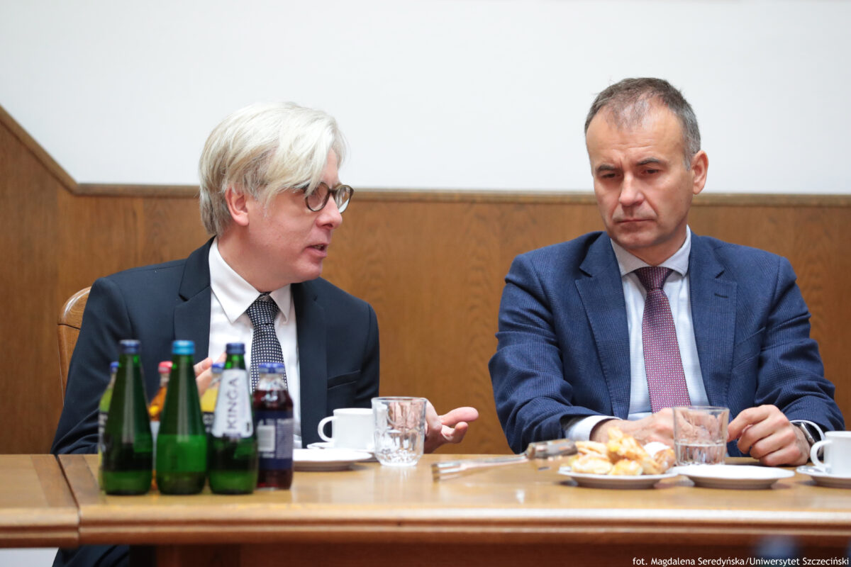 Dwóch mężczyzn w garniturach siedzących przy stole podczas spotkania. Po lewej stronie mężczyzna z jasnymi, siwymi włosami i okularami, w czarnym garniturze i białej koszuli, jest odwrócony w prawo i mówi do drugiego mężczyzny, gestykulując dłonią. Po prawej stronie mężczyzna z krótkimi, ciemnymi włosami, w niebieskim garniturze, białej koszuli i bordowym krawacie, ma poważny wyraz twarzy i trzyma dłonie blisko siebie na stole. Na stole stoją butelki (głównie zielone), szklanki, filiżanki oraz talerzyk z rogalikami lub drożdżówkami. W tle jest drewniana boazeria i jasna ściana. W prawym dolnym rogu widoczny jest napis: „fot. Magdalena Seredyńska/Uniwersytet Szczeciński”.
