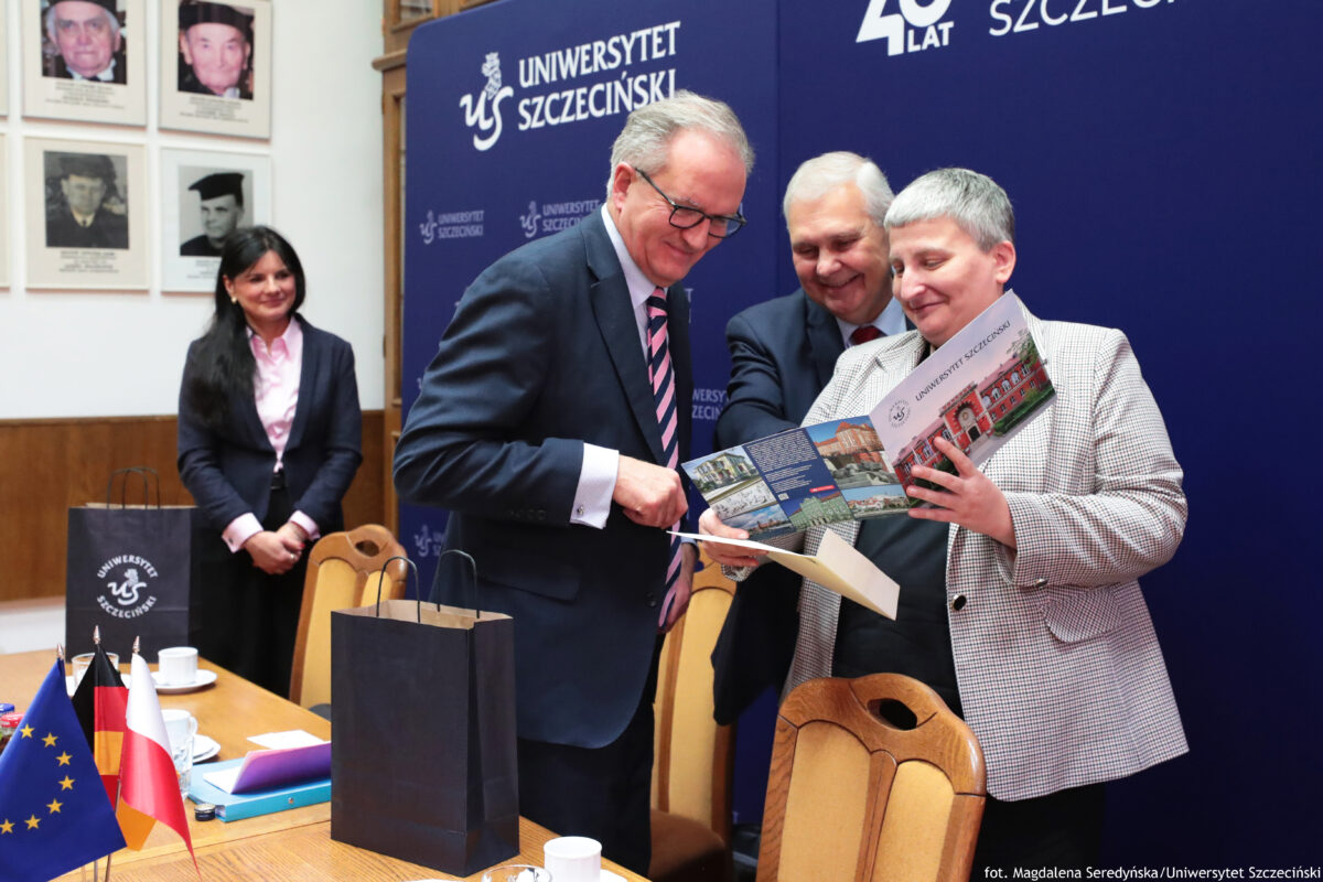 Scena z uroczystości na Uniwersytecie Szczecińskim, z trzema osobami na pierwszym planie i jedną osobą w tle. Trzy osoby stoją przy stole i uśmiechają się, oglądając dokument lub folder. Na pierwszym planie widać dwóch mężczyzn i jedną kobietę (wszyscy dorośli, jasna karnacja). Mężczyzna po lewej ma siwe włosy, okulary, ciemny garnitur i krawat w różowo-granatowe pasy; podaje lub pokazuje kartkę. Mężczyzna w środku ma siwe włosy i ciemny garnitur; stoi lekko z tyłu i też się uśmiecha. Kobieta po prawej ma krótkie siwe włosy i jasną marynarkę w drobną kratkę; trzyma otwarty, kolorowy folder z napisem „UNIWERSYTET SZCZECIŃSKI” i zdjęciami budynków. W tle stoi kobieta z długimi ciemnymi włosami, w ciemnym żakiecie i jasnoróżowej koszuli, obserwująca sytuację. Za osobami jest granatowa ścianka z białym napisem „UNIWERSYTET SZCZECIŃSKI” oraz „40 LAT”. Po lewej na ścianie wiszą oprawione portrety. Na stole stoją małe flagi (Unia Europejska, Niemcy i Polska), są też filiżanki, teczki/papierowe dokumenty i dwie ciemne torby prezentowe z logo uczelni. W prawym dolnym rogu jest podpis: „fot. Magdalena Seredyńska/Uniwersytet Szczeciński”.