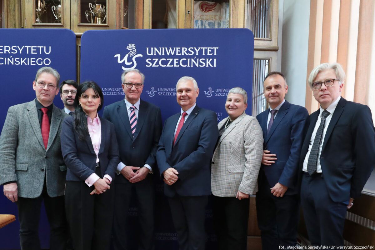 Grupa siedmiu dorosłych osób stojących w pomieszczeniu na tle niebieskiej ścianki z napisem „UNIWERSYTET SZCZECIŃSKI” i logo uczelni. Osoby są ubrane formalnie, głównie w garnitury i marynarki; część ma krawaty. W środku stoi mężczyzna w ciemnym garniturze z czerwonym krawatem; obok niego mężczyzna w okularach z krawatem w różowo-granatowe pasy oraz kobieta z długimi ciemnymi włosami w granatowej marynarce i jasnoróżowej koszuli. Po prawej stronie jest kobieta z krótkimi siwymi włosami w jasnej, kraciastej marynarce oraz dwóch mężczyzn w garniturach; skrajnie po prawej stoi mężczyzna o jasnych, siwych włosach w okularach, w czarnym garniturze i jasnym krawacie. Po lewej stronie jest mężczyzna w szarej marynarce i czerwonym krawacie, a za nim częściowo widoczny mężczyzna z ciemnymi włosami i brodą w okularach. Na dole po prawej widoczny jest podpis: „fot. Magdalena Seredyńska/Uniwersytet Szczeciński”.