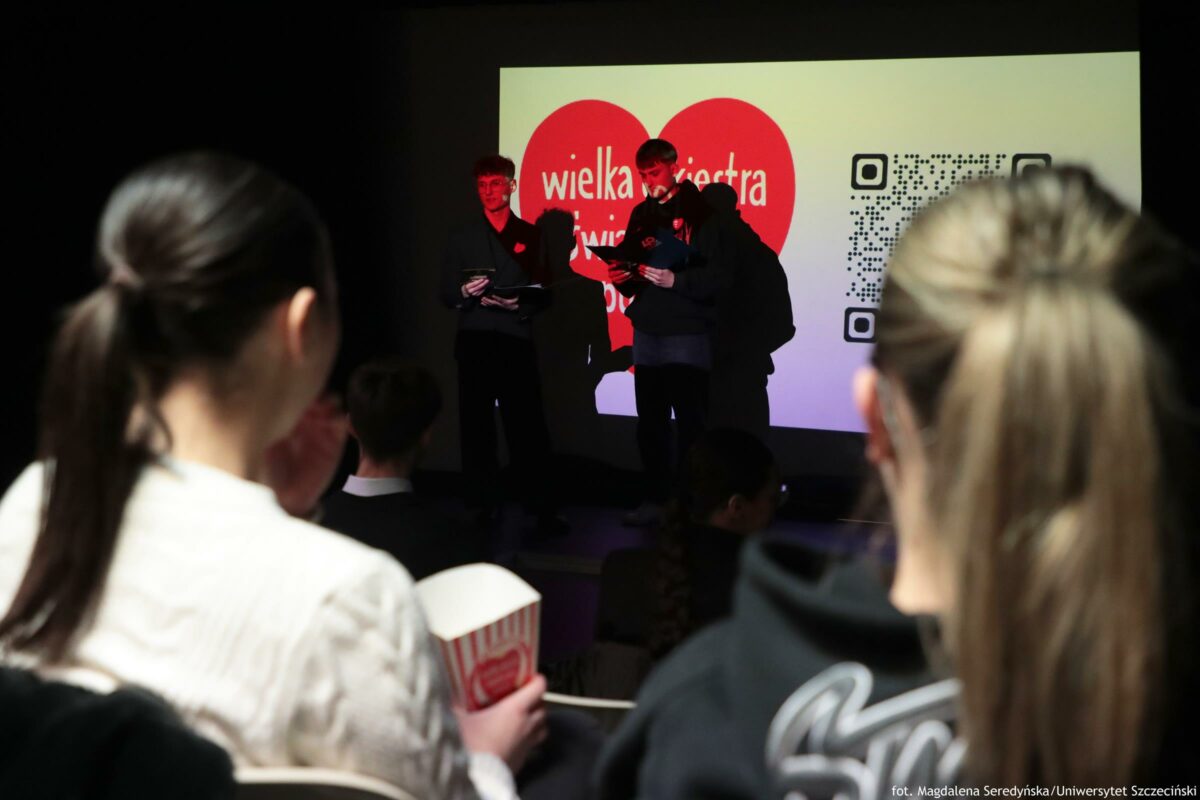 Sala z publicznością podczas wydarzenia na scenie. Na pierwszym planie widać tyły głów dwóch osób z długimi włosami związanymi w kucyk; jedna trzyma w ręku biało-czerwone pudełko, wyglądające jak na popcorn. W tle na scenie stoją trzy osoby w ciemnych ubraniach, dwie z nich trzymają kartki i wyglądają na czytające lub prowadzące. Za nimi jest duży ekran z czerwonym sercem i białym napisem „wielka orkiestra świątecznej pomocy”, a po prawej stronie ekranu widoczny jest duży kod QR. Na dole zdjęcia w prawym rogu jest podpis: „fot. Magdalena Seredyńska/Uniwersytet Szczeciński”.
