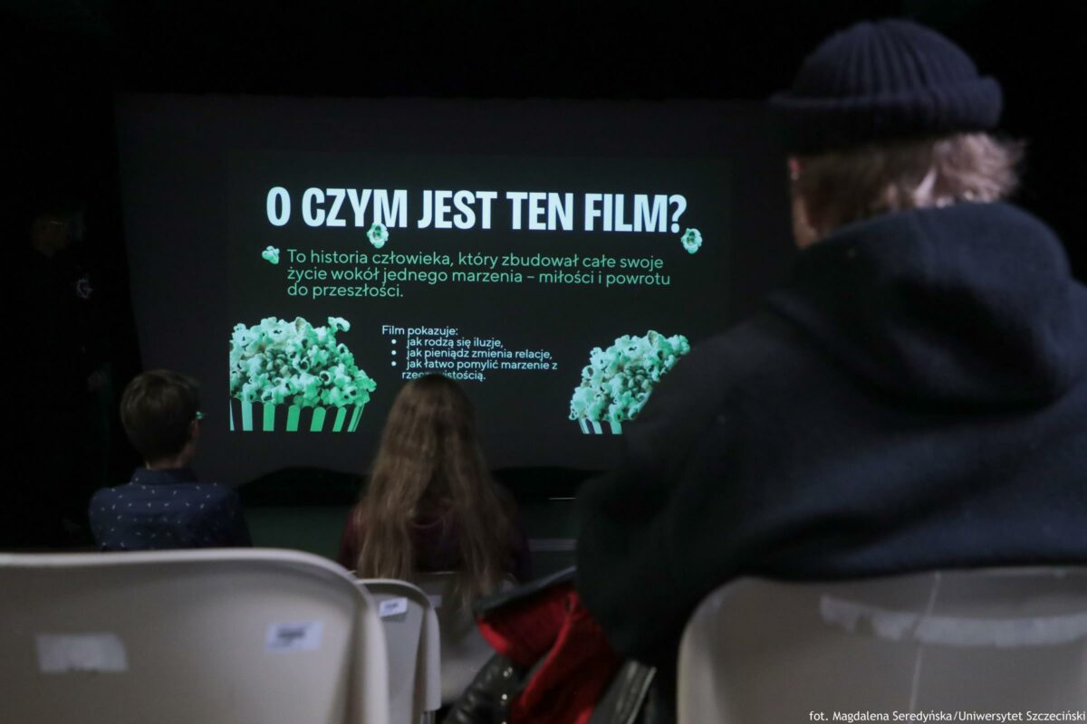 Ekran projekcyjny w ciemnej sali z dużym polskim napisem „O CZYM JEST TEN FILM?” oraz kilkoma linijkami tekstu poniżej i grafikami kubełków popcornu po obu stronach. Na pierwszym planie widać tyły głów kilku osób siedzących na jasnych krzesłach; po prawej zarys osoby w czapce, pośrodku osoba z długimi włosami, po lewej dziecko. Tekst na ekranie: O CZYM JEST TEN FILM? To historia człowieka, który zbudował całe swoje życie wokół jednego marzenia – miłości i powrotu do przeszłości. Film pokazuje: • jak rodzą się iluzje, • jak pieniądz zmienia relacje, • jak łatwo pomylić marzenie z rzeczywistością. fot. Magdalena Seredyńska/Uniwersytet Szczeciński