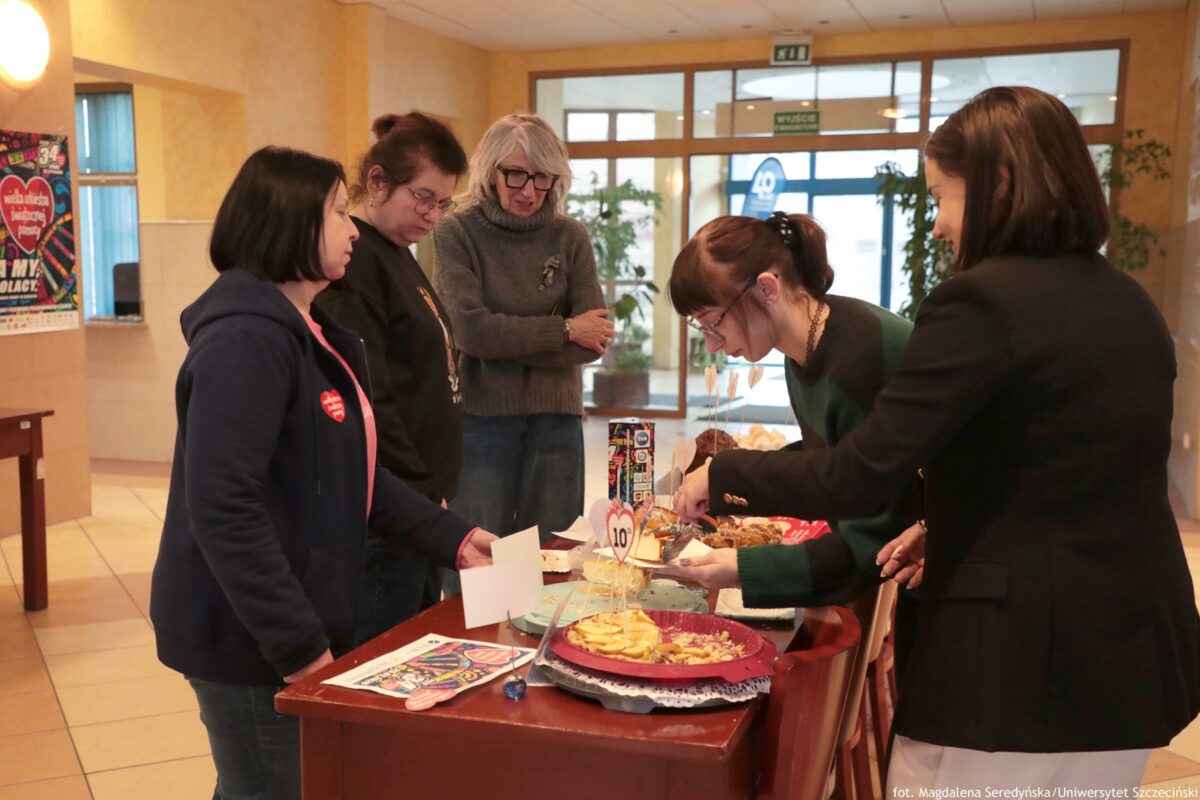 Grupa pięciu kobiet stojących przy stole z wypiekami w holu budynku. Dwie kobiety po prawej stronie pochylają się nad stołem i kroją lub nakładają jedzenie na talerzyk, a trzy pozostałe obserwują. Na stole leżą ciasta i inne wypieki na tacach, papierowe karteczki i widoczna okrągła kartka z liczbą „10” na patyczku. W tle są przeszklone drzwi i okna oraz zielone tabliczki „WYJŚCIE”. W prawym dolnym rogu jest podpis: „fot. Magdalena Seredyńska / Uniwersytet Szczeciński”.