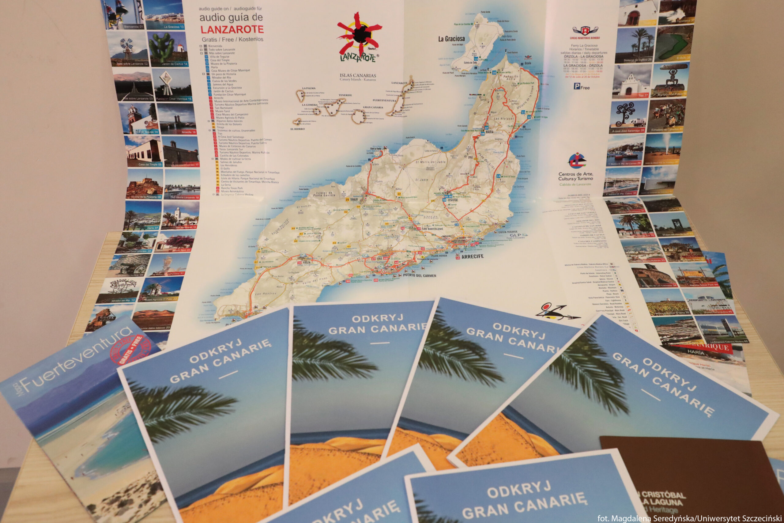Rozłożona mapa wyspy Lanzarote z dodatkowymi folderami turystycznymi na stole. Na środku leży duża, rozkładana mapa Lanzarote z zaznaczonymi drogami, miejscowościami i trasami w kolorze czerwonym. Po bokach dużej mapy znajdują się wąskie paski z wieloma małymi zdjęciami atrakcji turystycznych – krajobrazów, budynków, rzeźb i plaż. Na górze mapy widnieje napis „audio guia de LANZAROTE” z informacją „Gratis / Free / Kostenlos” oraz logo z czerwonym kształtem przypominającym słońce i czarną sylwetką. Na dole obrazu rozłożone są liczne foldery w języku polskim z błękitną okładką i zdjęciem plaży oraz wydm; na każdym widać biały napis „ODKRYJ GRAN CANARIĘ”. Po lewej stronie leży inny folder z napisem „Fuerteventura” oraz zdjęciem jasnej plaży i turkusowego morza. W prawym dolnym rogu częściowo widoczne są brązowe broszury z białymi napisami, sugerujące inne informacje turystyczne.