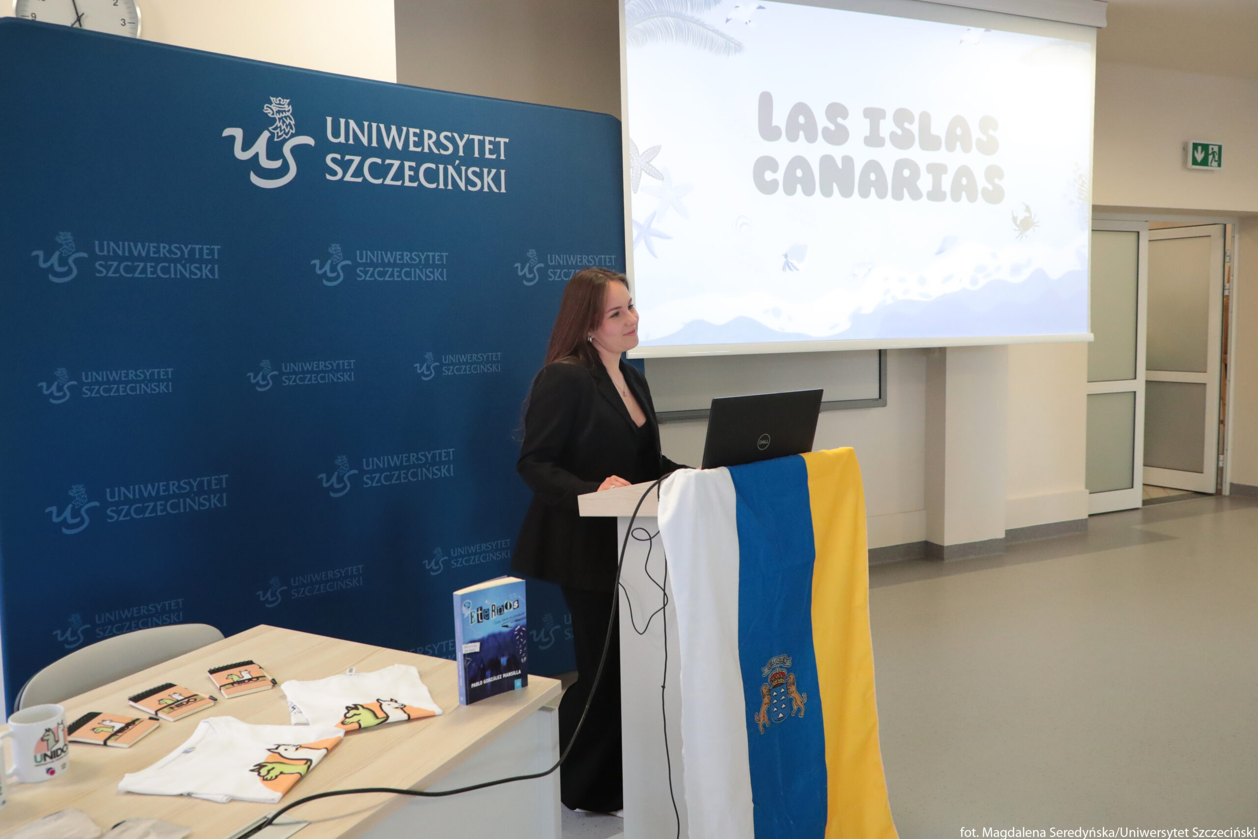 Kobieta stojąca za mównicą podczas prezentacji na uczelni. Kobieta ma jasną skórę, długie ciemne włosy i ubrana jest w czarny garnitur. Stoi przy białej mównicy, na której leży rozwieszona flaga w pionowe pasy: biały, niebieski i żółty, z herbem pośrodku niebieskiego pasa. Na mównicy stoi otwarty czarny laptop, z którego wychodzi czarny kabel. Za kobietą po lewej stronie znajduje się duża niebieska ścianka z powtarzającym się białym napisem „UNIWERSYTET SZCZECIŃSKI” oraz białym logo z literami „US”. W lewym górnym rogu na ścianie jest okrągły zegar. Z prawej strony, nad głową kobiety, znajduje się rozwinięty ekran projekcyjny. Na ekranie widoczny jest jasny slajd z napisem „LAS ISLAS CANARIAS” w dużych, zaokrąglonych literach, z rysunkami fal, ptaków i elementów morskich. Przed mównicą stoi jasne biurko. Na blacie leżą dwie białe koszulki z kolorowym graficznym motywem, kilka prostokątnych przedmiotów przypominających notesy lub broszury, biała porcelanowa filiżanka z nadrukiem „UNI” i logo, oraz stojąca książka z niebieską okładką. W tle po prawej stronie widoczne są drzwi z przeszklonymi panelami oraz zielony znak wyjścia ewakuacyjnego nad drzwiami.