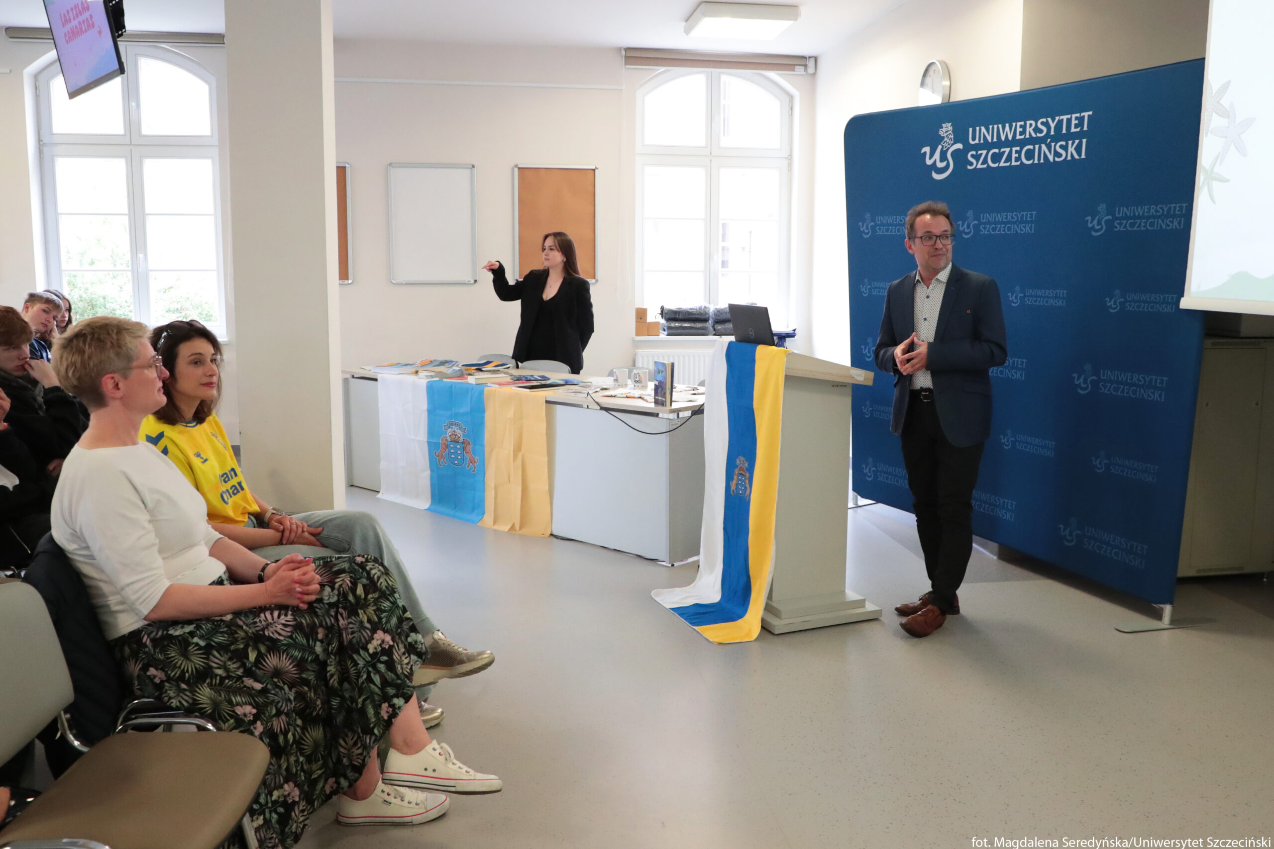 Sala wykładowa na Uniwersytecie Szczecińskim podczas spotkania lub prezentacji. Po prawej stronie stoi mężczyzna w ciemnej marynarce, jasnej koszuli i ciemnych spodniach, oparty o jasną mównicę. Na mównicy zwisa pionowo flaga w trzech pasach: białym, niebieskim i żółtym, z herbem w środkowej części. Za mężczyzną znajduje się duża, niebieska ścianka z białym napisem „UNIWERSYTET SZCZECIŃSKI” i powtarzającym się logo uczelni. Po lewej stronie kadru siedzi kilka osób na krzesłach, w pierwszym rzędzie kobieta w białej bluzce i długiej, ciemnej, kwiecistej spódnicy oraz obok młoda kobieta w żółtej koszulce sportowej z niebieskimi nadrukami. Obie mają jasną skórę. Dalej siedzą kolejne osoby, częściowo zasłonięte. W głębi sali, za długim stołem, stoi kobieta w czarnym ubraniu, wykonująca gesty rękami, prawdopodobnie tłumacząca na polski język migowy. Na stole leżą rozłożone materiały: broszury, książki i inne drukowane przedmioty. Z przodu stołu przewieszona jest poziomo flaga w trzech pionowych pasach: białym, niebieskim i żółtym, z herbem pośrodku niebieskiego pasa, obok niej jasny, jednokolorowy materiał. Na końcu stołu stoi otwarty laptop. W tle widoczne są wysokie, jasne okna z białymi ramami oraz puste tablice na ścianie. W lewym górnym rogu, przy suficie, wisi ekran lub monitor z czerwonym napisem, którego treść jest zbyt mała, by ją odczytać.