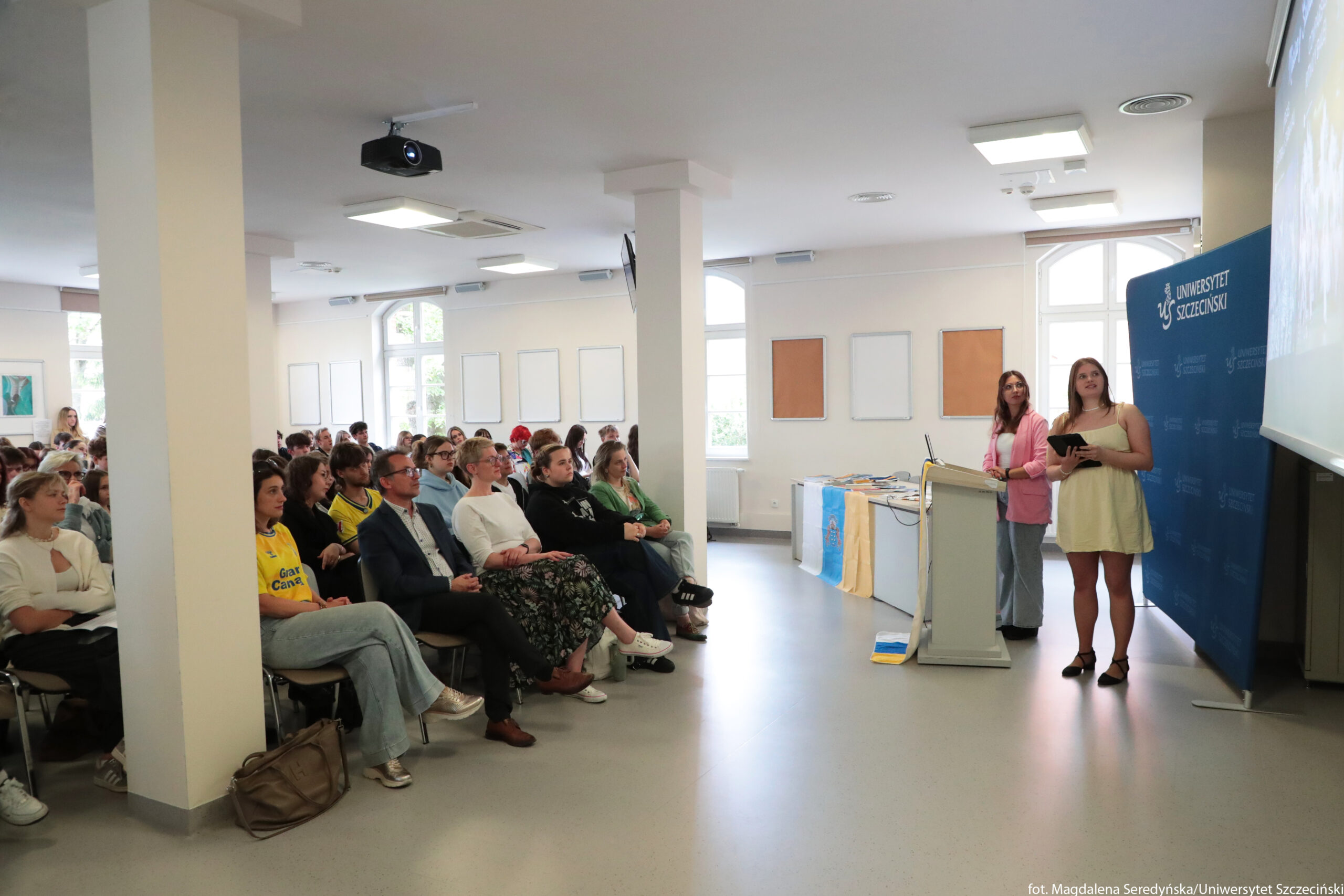Sala wykładowa z publicznością i dwiema prelegentkami przy mównicy. Po lewej stronie siedzi kilkadziesiąt osób w kilku rzędach krzeseł; większość to młode osoby, ale widać też kilka starszych, wszyscy skupieni na wystąpieniu. W pierwszym rzędzie siedzi mężczyzna w garniturze oraz osoby w swobodnych ubraniach, między innymi kobieta w żółtej koszulce sportowej i kobieta w długiej, wzorzystej spódnicy. Po prawej stronie, przy jasnej mównicy z laptopem, stoją dwie kobiety: jedna w krótkiej, jasnokremowej sukience trzyma tablet, druga w różowej marynarce i jasnych spodniach stoi bliżej tła. Za nimi znajduje się niebieska ścianka z białym logo i napisem „UNIWERSYTET SZCZECIŃSKI”. Na stole obok mównicy leżą rozłożone materiały i kilka flag lub szali w kolorach niebieskim i żółtym. Na suficie widać projektor, a na ścianach prostokątne ramy lub tablice; przez okna wpada dzienne światło.