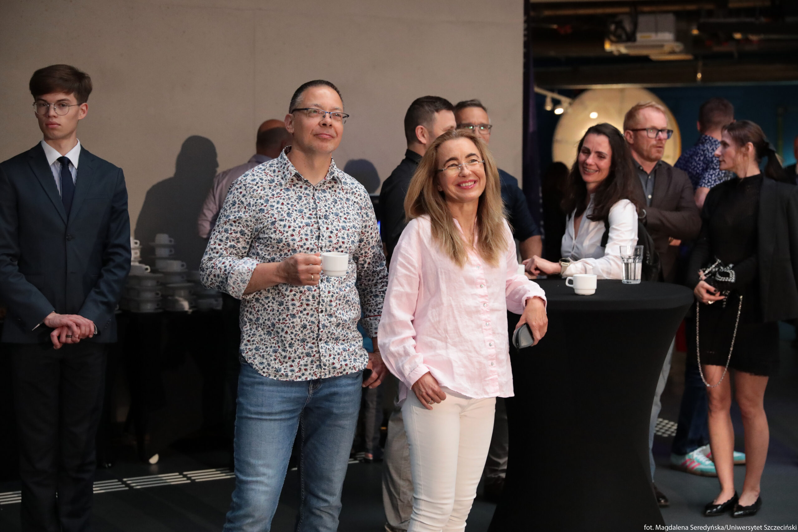 Grupa osób podczas spotkania towarzyskiego lub konferencyjnego w pomieszczeniu. Na pierwszym planie po lewej stronie stoi mężczyzna o jasnej karnacji, z krótkimi ciemnymi włosami i okularami, ubrany w koszulę w drobny, kolorowy wzór i niebieskie dżinsy; trzyma w dłoni białą filiżankę. Obok niego, po prawej stronie, stoi kobieta o jasnej karnacji i długich, rozpuszczonych blond włosach, w jasnoróżowej lnianej koszuli i jasnych spodniach; opiera rękę o wysoki, okrągły stolik koktajlowy nakryty czarnym materiałem i uśmiecha się. Za nimi zgromadzonych jest kilka osób rozmawiających w niewielkich grupkach; część trzyma szklanki lub filiżanki, część ma założone ręce. Po lewej stronie kadru, osobno, stoi młody mężczyzna o jasnej karnacji, w ciemnym garniturze z krawatem i okularach, z dłońmi złożonymi przed sobą. W głębi, przy czarnym bufecie, ustawione są stosy białych spodków i filiżanek. Tło po prawej stronie jest ciemniejsze, z elementami oświetlenia sufitowego i nieostrymi postaciami uczestników wydarzenia.