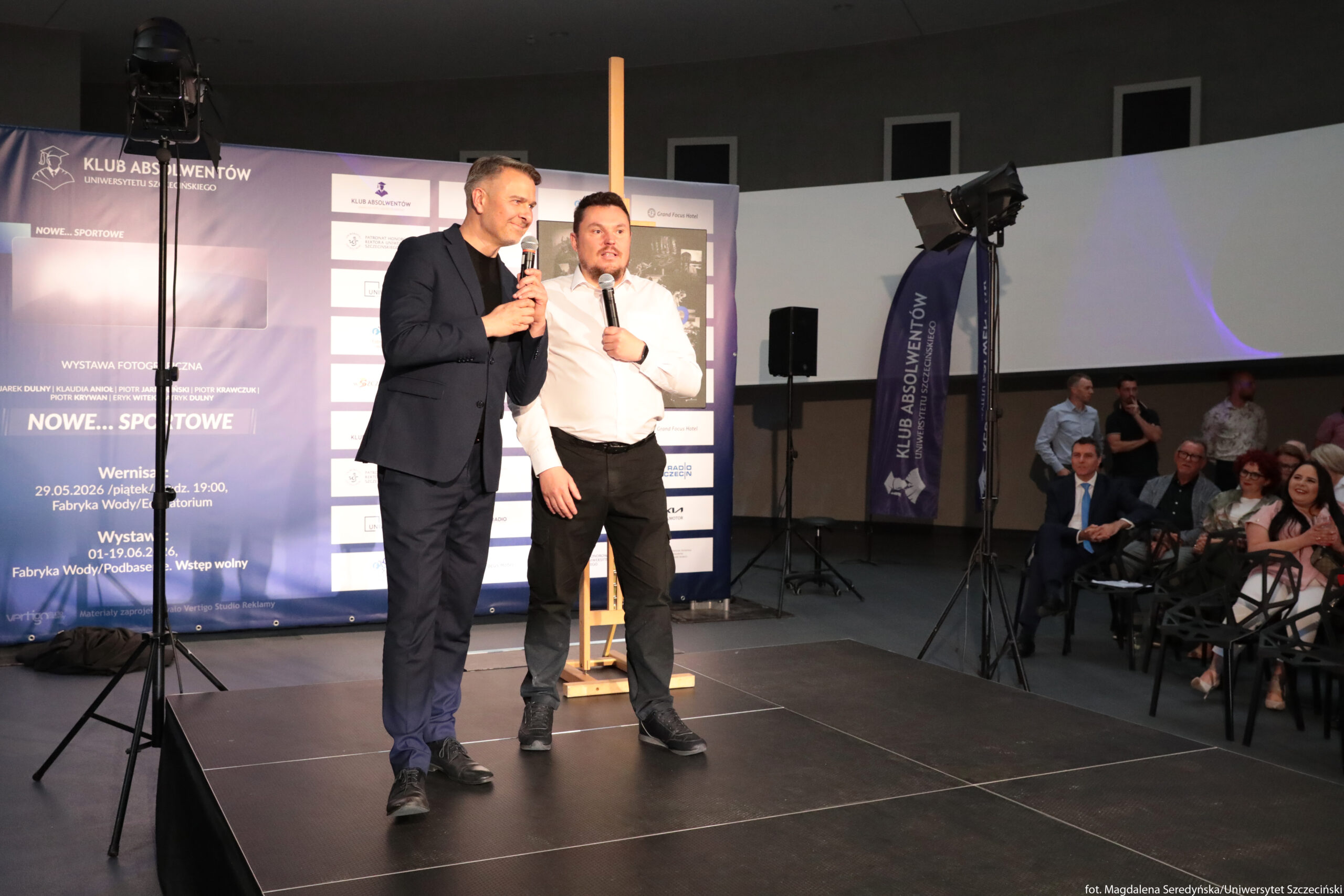 Dwóch mężczyzn stojących na podwyższonej scenie w sali wydarzeń. Obaj trzymają mikrofony i są oświetleni jasnym światłem z reflektorów ustawionych przed sceną. Mężczyzna po lewej ma krótko przycięte siwiejące włosy, ciemny garnitur, czarną koszulkę i ciemne buty, uśmiecha się i patrzy w stronę publiczności. Mężczyzna po prawej ma krótkie, ciemne włosy, białą koszulę z podwiniętymi rękawami, ciemne spodnie i sportowe buty; stoi blisko towarzysza, jedną rękę ma opuszczoną, drugą trzyma mikrofon przy ustach. Za nimi znajduje się duży, niebieski baner z napisem „KLUB ABSOLWENTÓW UNIWERSYTETU SZCZECIŃSKIEGO”, logotypami partnerów oraz informacjami o wystawie fotograficznej „NOWE… SPORTOWE”; na banerze czytelne są fragmenty tekstu „WYSTAWA FOTOGRAFICZNA”, „Wernisaż 29.05.2026 /piątek/ godz. 19.00, Fabryka Wody/Audytorium” oraz „Wystawa 01–19.06.26, Fabryka Wody/Podbasenie. Wstęp wolny”. Po prawej stronie widać pionową niebieską flagę z napisem „KLUB ABSOLWENTÓW UNIWERSYTETU SZCZECIŃSKIEGO”. Przed sceną, po prawej stronie kadru, siedzi grupa osób na krzesłach; część z nich klaszcze, jedna kobieta w jasnej sukience szeroko się uśmiecha. Na dalszym planie stoją osoby rozmawiające, a przy ścianach ustawione są kolumny głośnikowe i sprzęt oświetleniowy.