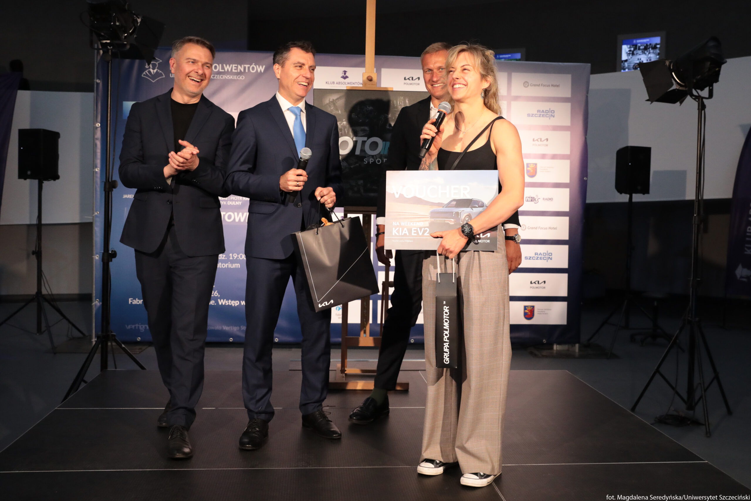 Grupa pięciu dorosłych osób na scenie podczas uroczystości wręczenia nagrody. Trzech mężczyzn po lewej stronie ma na sobie eleganckie granatowe garnitury, białe koszule i krawaty; wszyscy uśmiechają się, dwóch trzyma mikrofony, jeden klaszcze. Po prawej stoi kobieta o jasnych, krótkich, lekko rozczochranych włosach, jasnej karnacji i atletycznej sylwetce, ubrana w czarny top na ramiączkach, luźne beżowe spodnie i sportowe buty. Kobieta trzyma w jednej ręce mikrofon i duży prostokątny karton z napisem „VOUCHER na weekend z KIA EV2” oraz zdjęciem samochodu, w drugiej ręce podłużną torbę prezentową z nadrukiem „GRUPA POLMOTOR”. Przed nią, nieco niżej, widać jeszcze jedną czarną torbę prezentową z ukośnym jasnym wzorem i małym logo KIA, trzymaną przez mężczyznę w garniturze. W tle stoją ścianki sponsorskie z licznymi logotypami i tekstem w języku polskim, m.in. „KLUB ABSOLWENTÓW”, „UNIWERSYTET SZCZECIŃSKI”, „KIA”, „Radio Szczecin”, a także duże reflektory oświetlające scenę.