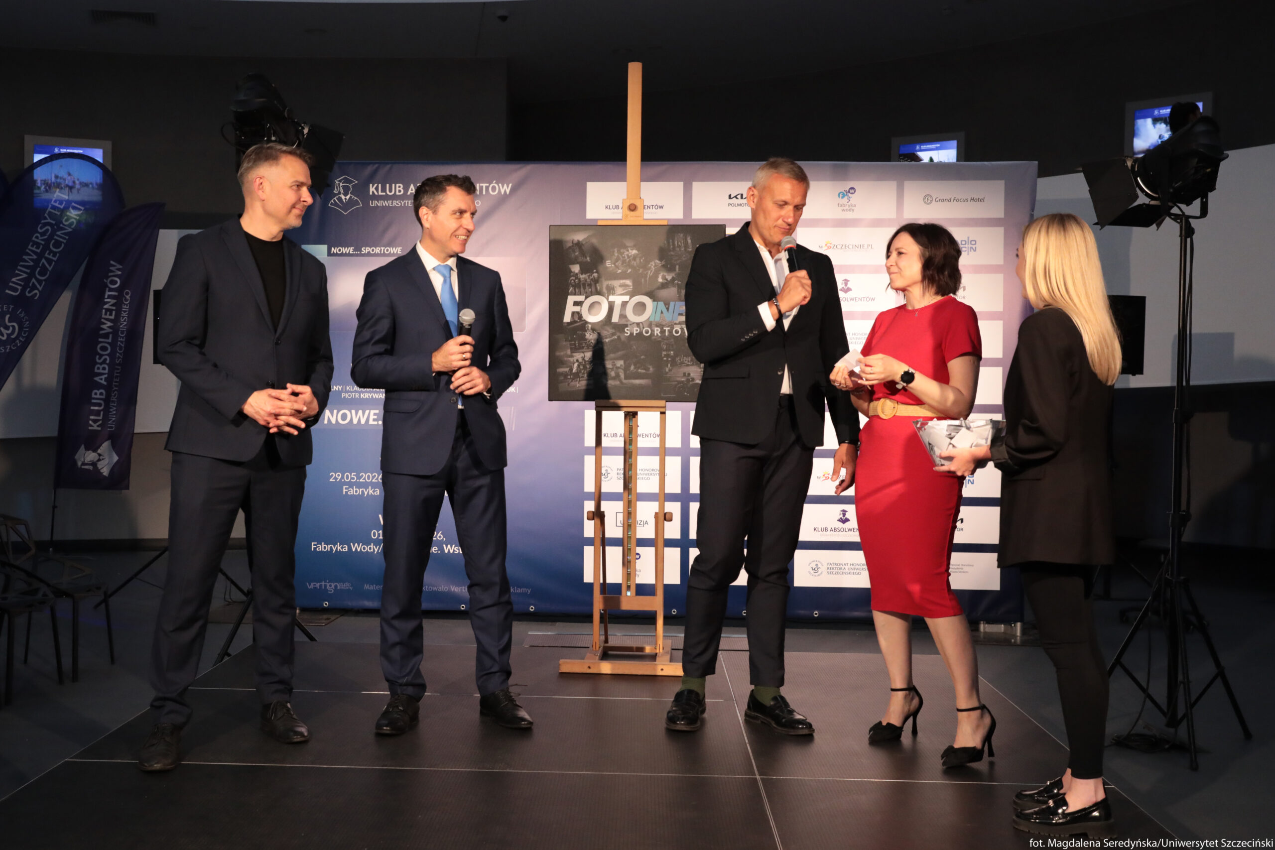 Pięć osób stojących na scenie podczas oficjalnego wydarzenia. Po lewej stronie stoi mężczyzna w granatowym garniturze bez krawata, z krótkimi, siwiejącymi włosami, trzyma dłonie splecione przed sobą i uśmiecha się w stronę środka. Obok niego drugi mężczyzna w ciemnym garniturze z jasnoniebieskim krawatem, trzyma w prawej ręce mikrofon i spogląda w prawo. Na środku stoi wysoki mężczyzna w czarnym garniturze z białą koszulą, również z mikrofonem, lekko pochylony w stronę kobiety, z którą rozmawia. Kobieta obok ma średniej długości ciemne włosy, ma na sobie dopasowaną, ciemnoczerwoną sukienkę do połowy łydki z paskiem w talii oraz czarne buty na obcasie; w dłoniach trzyma niewielkie pudełko lub upominek. Po prawej stronie stoi kobieta z długimi, jasnymi włosami, ubrana w czarną marynarkę i spodnie, trzyma w rękach pudełko lub torebkę prezentową, jest zwrócona w stronę grupy. Za osobami znajduje się rozstawiona na sztaludze plansza z napisem „FOTONEW… SPORTOWE” (środkowy fragment jest zasłonięty światłem). W tle widać duży niebiesko-biały baner z logotypami i napisami, między innymi „KLUB ABSOLWENTÓW” oraz „UNIWERSYTETU SZCZECIŃSKIEGO”, a po lewej stronie dwie pionowe flagi z podobnymi napisami. Po bokach ustawione są statywy z dużymi lampami studyjnymi, co sugeruje, że jest to zorganizowane wydarzenie z oświetleniem scenicznym.