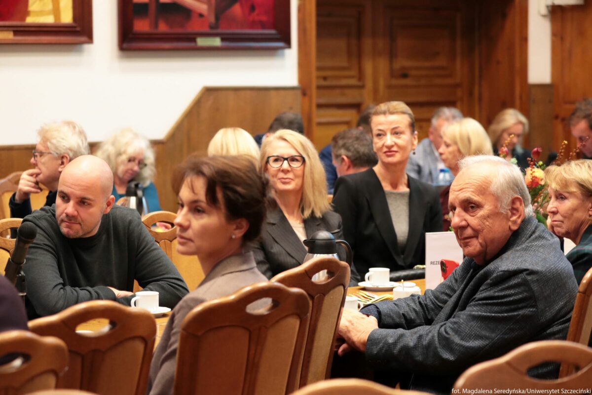 Sala Senatu US z grupą osób siedzących przy drewnianych stołach. Przy przednim stole siedzi kilka osób: po lewej stronie mężczyzna z ogoloną głową w ciemnym swetrze, obok niego kobieta z ciemnymi włosami upiętymi z tyłu w jasnym żakiecie. Nieco dalej, w środku kadru, siedzi kobieta z prostymi blond włosami do ramion, w ciemnej marynarce i dużych czarnych okularach. Po jej prawej stronie znajduje się kobieta z jasnymi, zaczesanymi do tyłu włosami, w czarnej marynarce i szarym topie, patrząca lekko w bok z delikatnym uśmiechem. Najbliżej prawej krawędzi zdjęcia siedzi starszy mężczyzna z siwymi włosami, w niebieskiej marynarce, zwrócony nieco w stronę obiektywu. Przy stołach stoją porcelanowe filiżanki na spodkach, talerzyki i termos, a na jednym ze stołów kartka z napisem „REZERWACJA”. W tle widać więcej osób siedzących przy kolejnych stołach oraz drewniane ściany i drzwi; wyżej wiszą duże obrazy w ciemnych ramach.