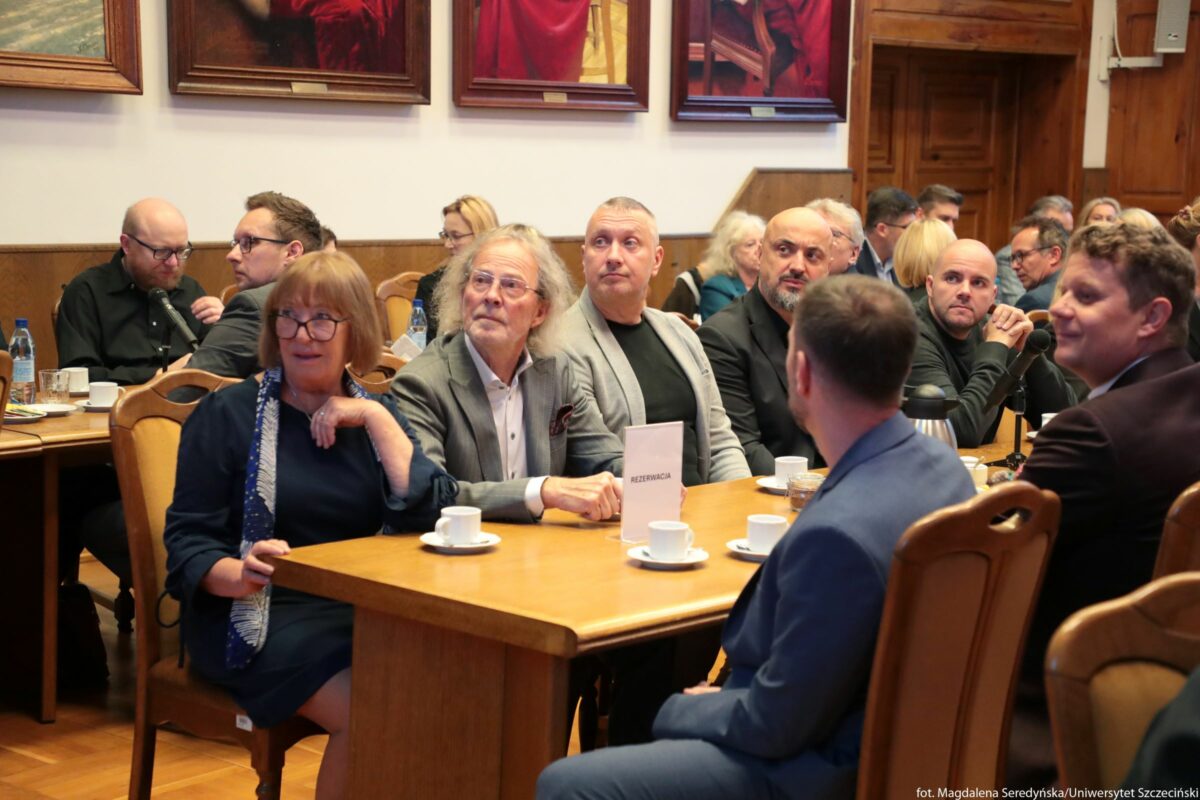 Sala Senatu US z drewnianymi krzesłami i stołami, przy jednym z nich grupa osób w eleganckich strojach siedząca przy kawie. Na pierwszym planie znajduje się prostokątny drewniany stół, na którym stoją białe filiżanki na spodkach, dzbanek termiczny z napojem i kartonik z napisem „REZERWACJA”. Przy tym stole siedzi kilka osób: z lewej strony kobieta w granatowej sukience i szalu, w okularach, z krótkimi jasnobrązowymi włosami; obok niej mężczyzna z siwymi, dłuższymi włosami i brodą, w szarej marynarce i białej koszuli, w okularach; dalej mężczyzna o krótko ostrzyżonych, jasnych włosach, w jasnej marynarce i czarnym T‑shircie; obok niego mężczyzna łysy, z krótką brodą, ubrany na czarno. Tyłem do aparatu siedzi mężczyzna w niebieskim garniturze, a po prawej stronie kadru widać mężczyznę w ciemnej marynarce, uśmiechniętego, lekko odwróconego w stronę środka stołu. W głębi pomieszczenia przy innych stołach siedzą kolejne osoby, ubrane oficjalnie, rozmawiające lub słuchające, na stołach stoją butelki wody, filiżanki i naczynia. Ściany sali są wyłożone drewnem w dolnej części, a wyżej wiszą duże obrazy w ciemnych ramach przedstawiające postacie ubrane w czerwone szaty. Z tyłu po prawej stronie widoczne są drewniane drzwi. W prawym dolnym rogu zdjęcia znajduje się drobny napis: „fot. Magdalena Seredyńska/Uniwersytet Szczeciński”.